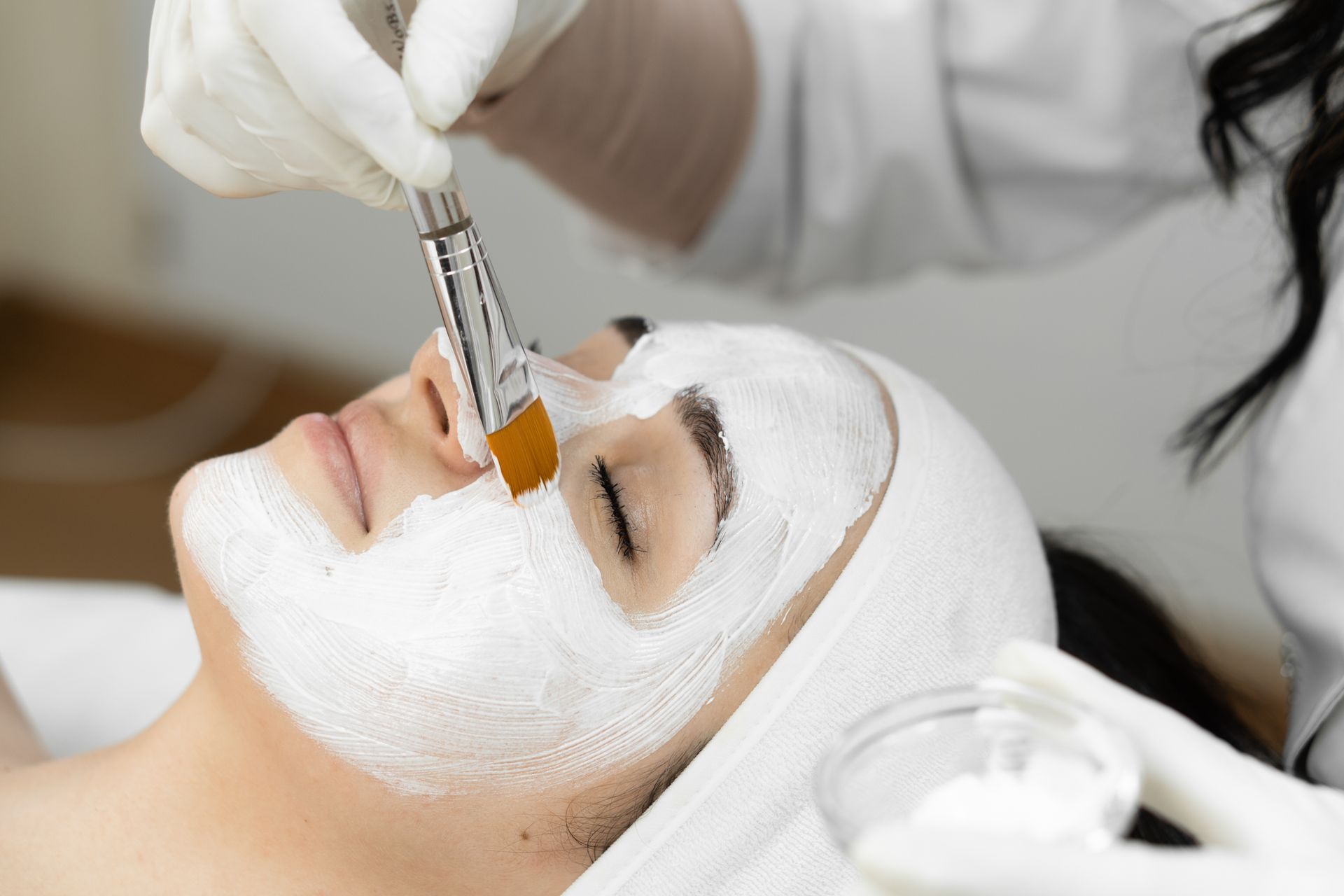 Facial mask applied with a brush to a person's face. White mask, gloved hands, light setting.
