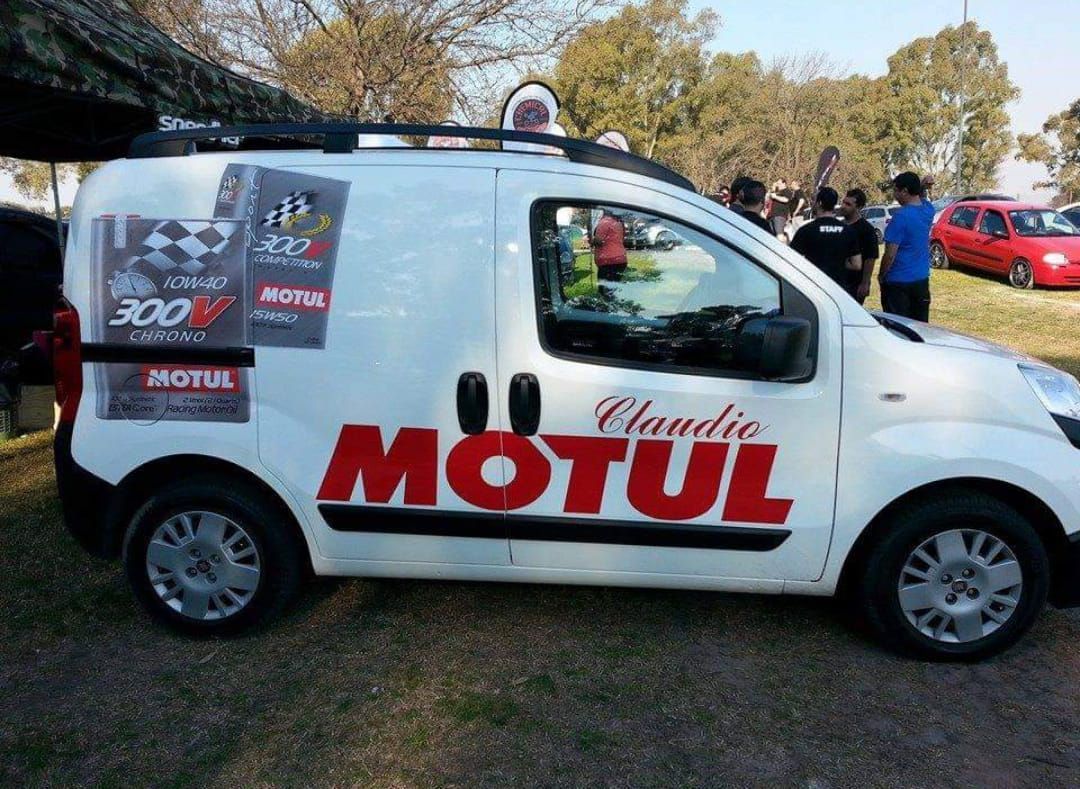 Furgoneta blanca con la marca Motul estacionada sobre el césped, con gente al fondo.