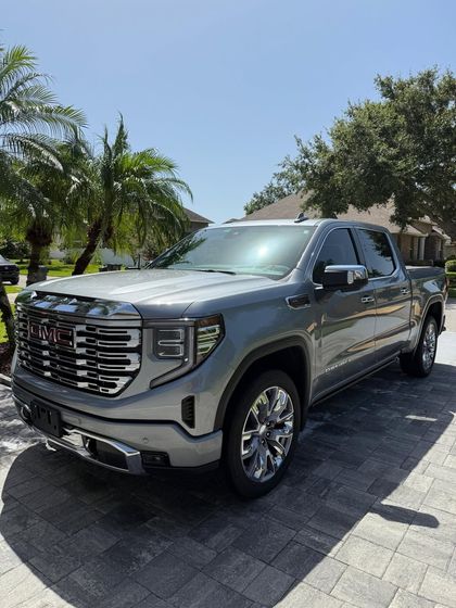 Close-up of a gray car hood with sparkly paint and a black grill; parked in front of a garage.