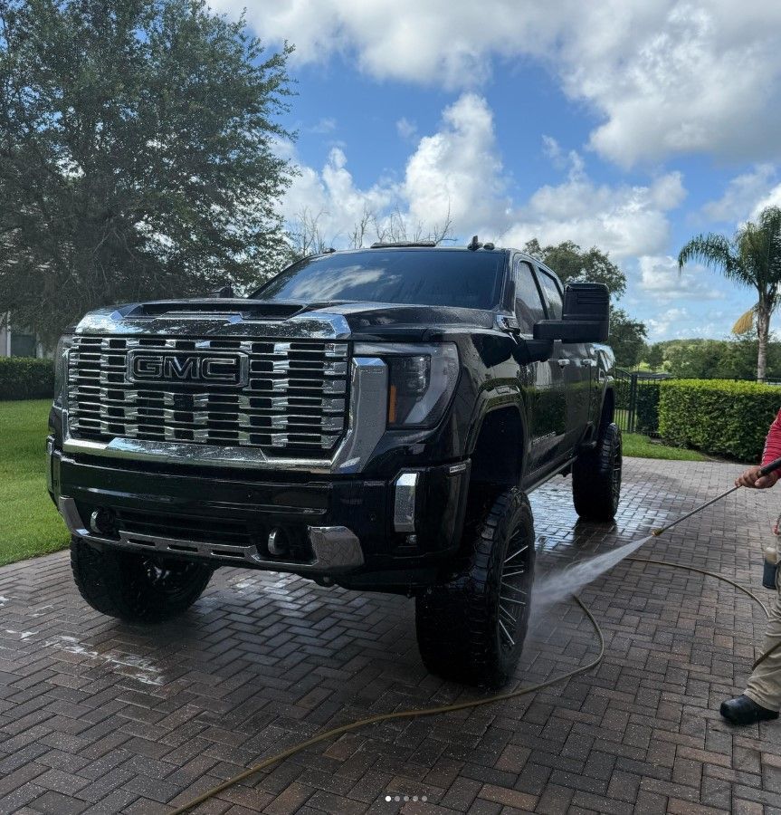 Man washing a blue car with a brush and hose outside on a sunny day.