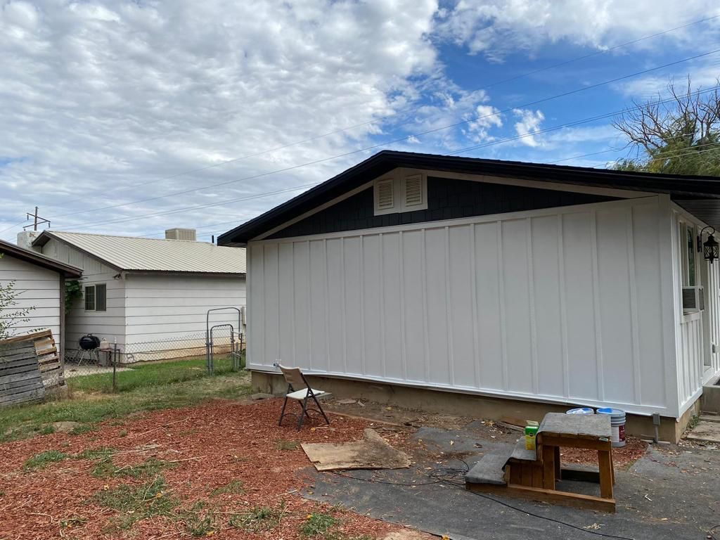 A man in a green shirt paints a house exterior with a spray gun, transitioning from red to white siding on a sunny day.