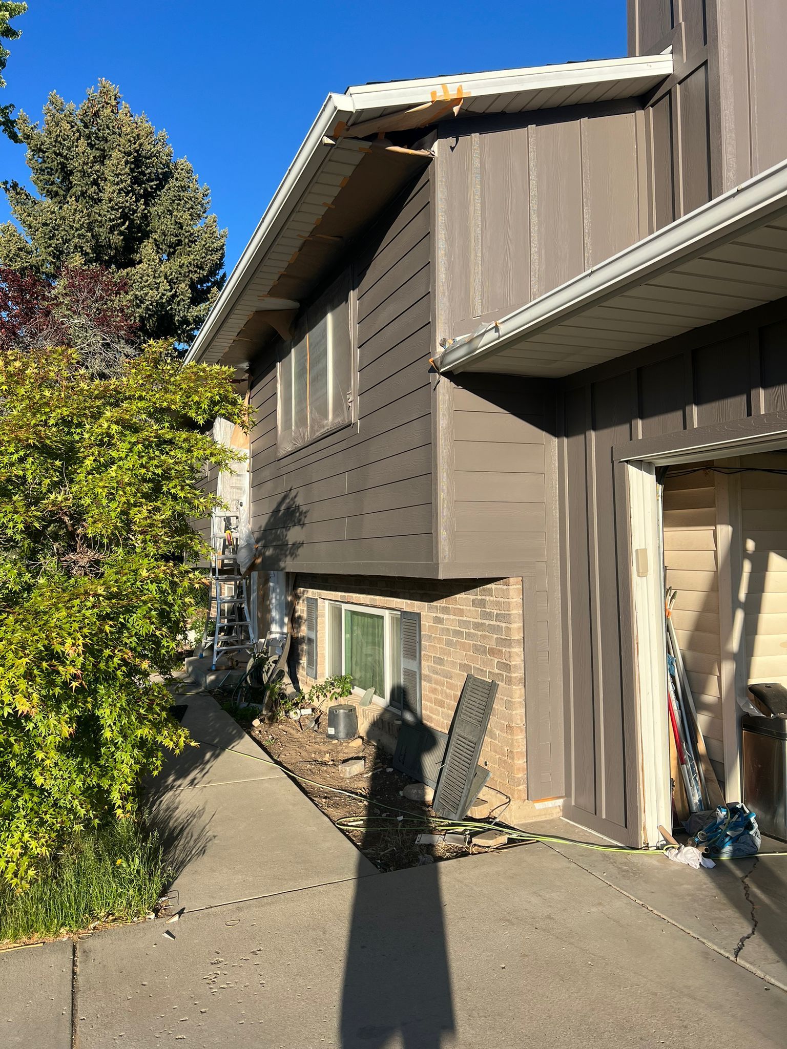 A man in jeans and a gray t-shirt uses a paint sprayer on a house exterior, with a paint supply canister on the ground.