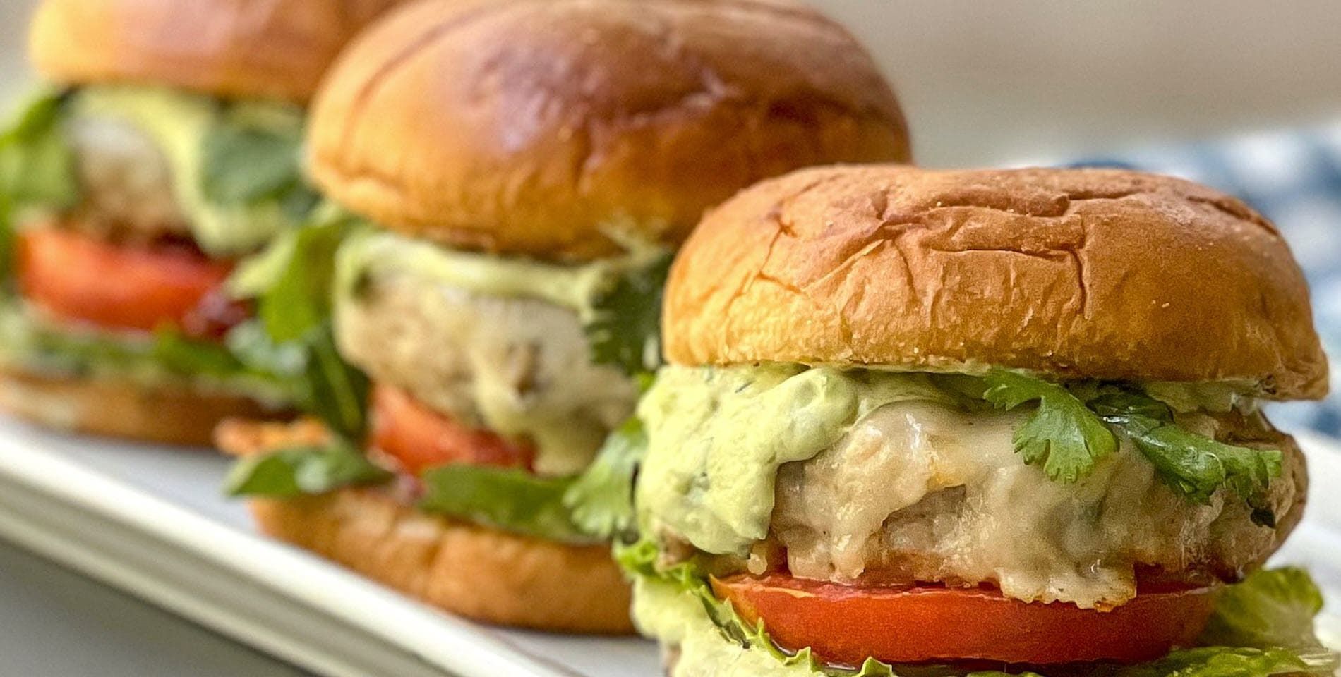 Three burgers on a white platter: Chicken patties with melted cheese, tomato, lettuce, and avocado crema on toasted buns.