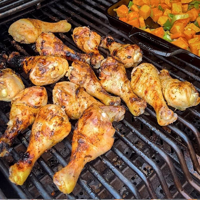 Chicken legs are cooking on a grill next to a pan of vegetables