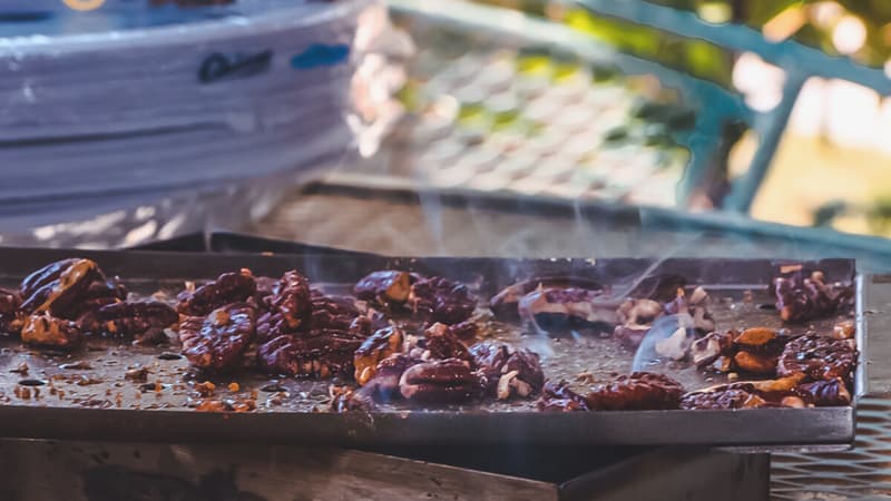 Candied pecans smoking on a metal tray over an outdoor grill.