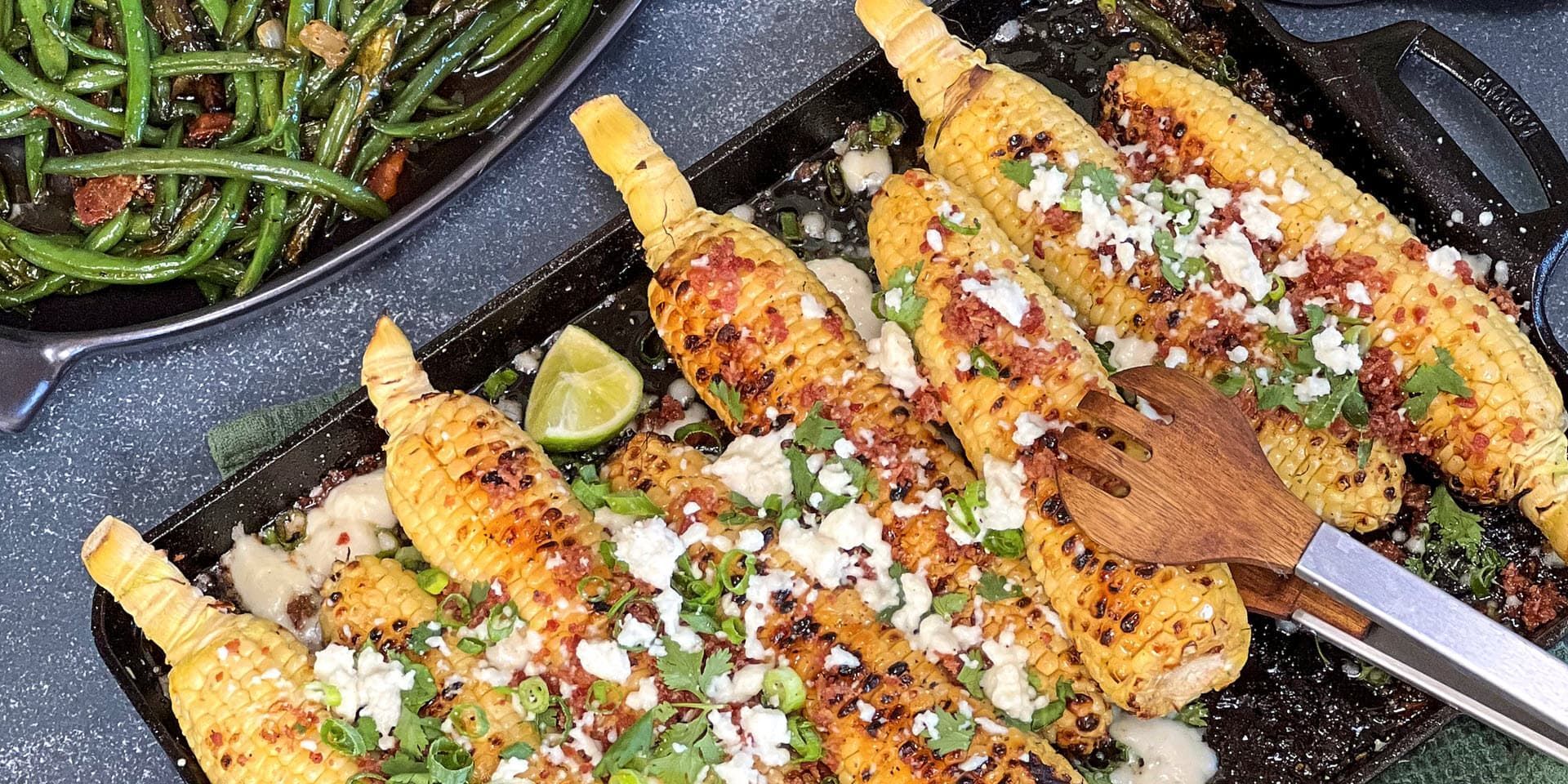 A tray of corn on the cob with a spatula and green beans on a table.