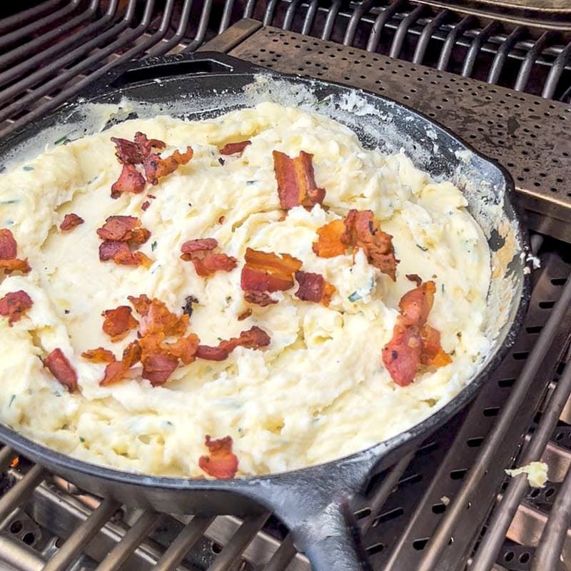 mashed potatoes with chives sprinkled with bacon bits in cast iron pan on grill