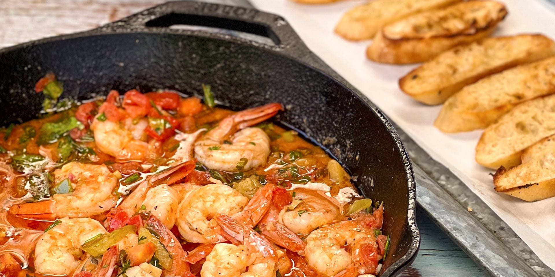 A skillet filled with seasoned & grilled shrimp next to slices of baguette bread