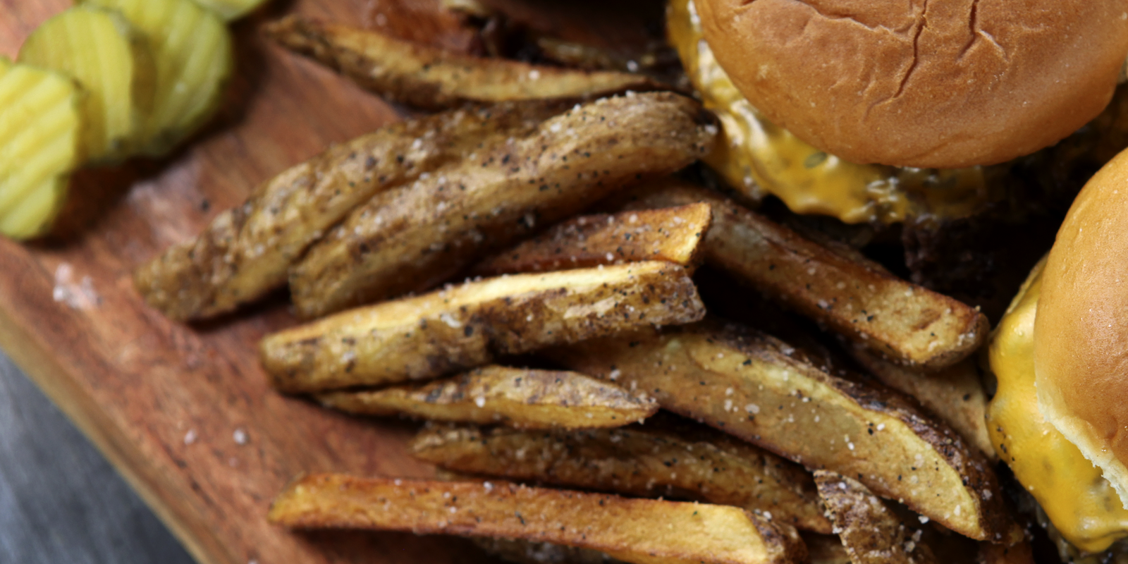 Close-up of seasoned fries, pickles, and cheeseburgers on a wooden serving board.
