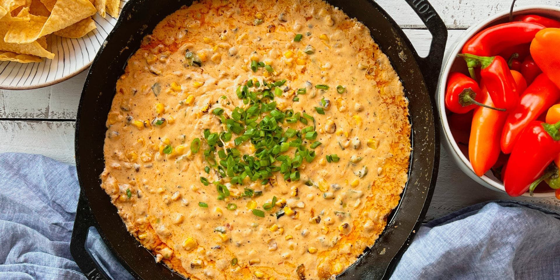 A cast iron skillet of cheesy corn dip, topped with green onions, beside chips and peppers.