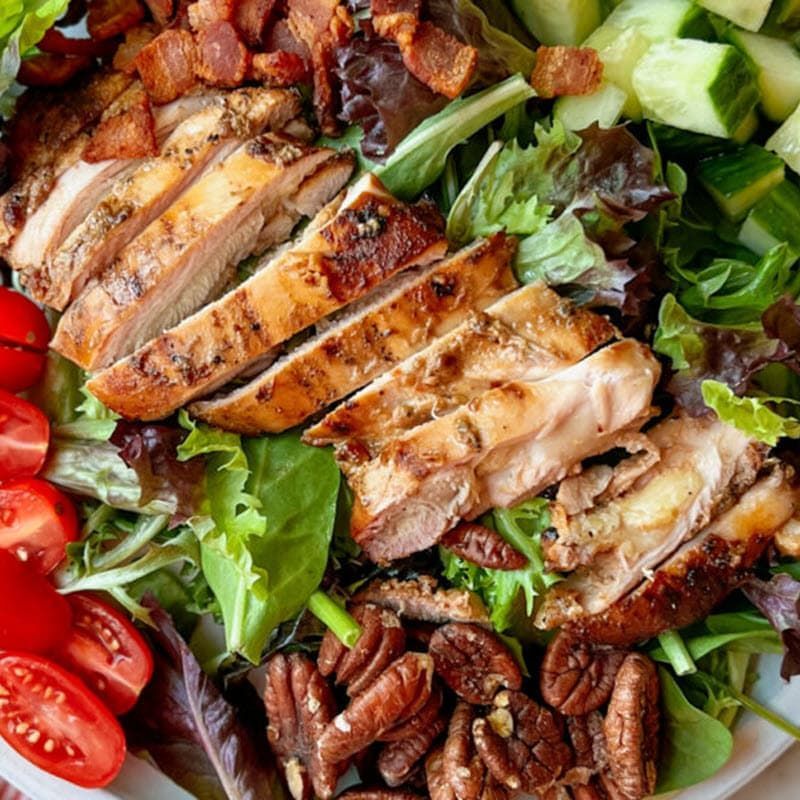 Sliced grilled chicken thighs on bed of lettuce with cucumber tomato and walnuts