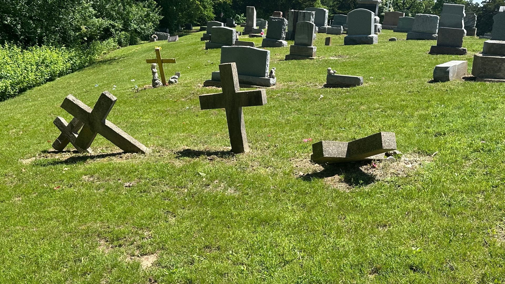 Grassy cemetery with several stone cross headstones, some leaning or fallen over, on a sunny day.  By Mary Berry - Art
