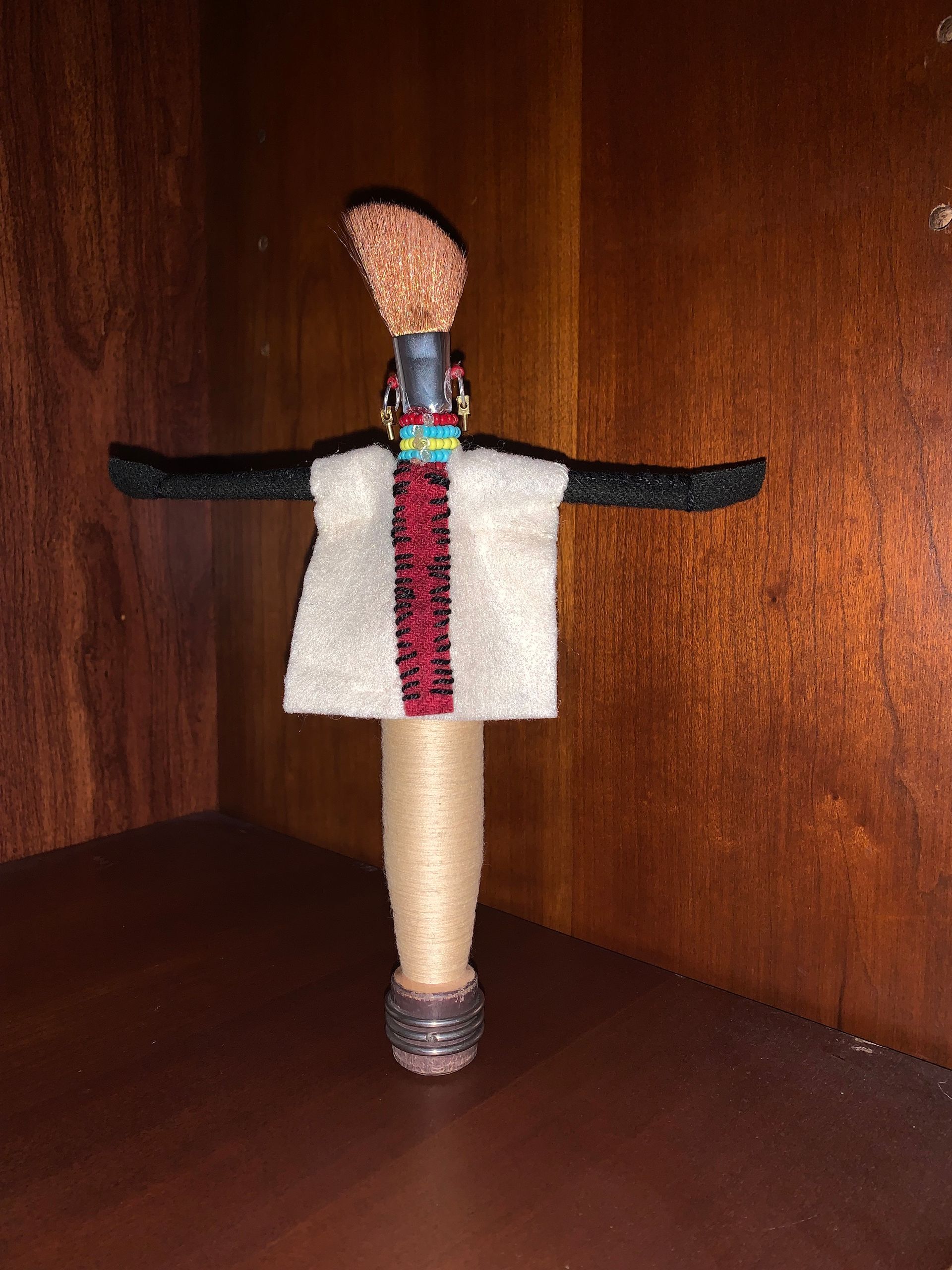 Handmade doll made from a paintbrush, with a felt shirt and black arms; set on a brown shelf.  By Mary Berry - Art