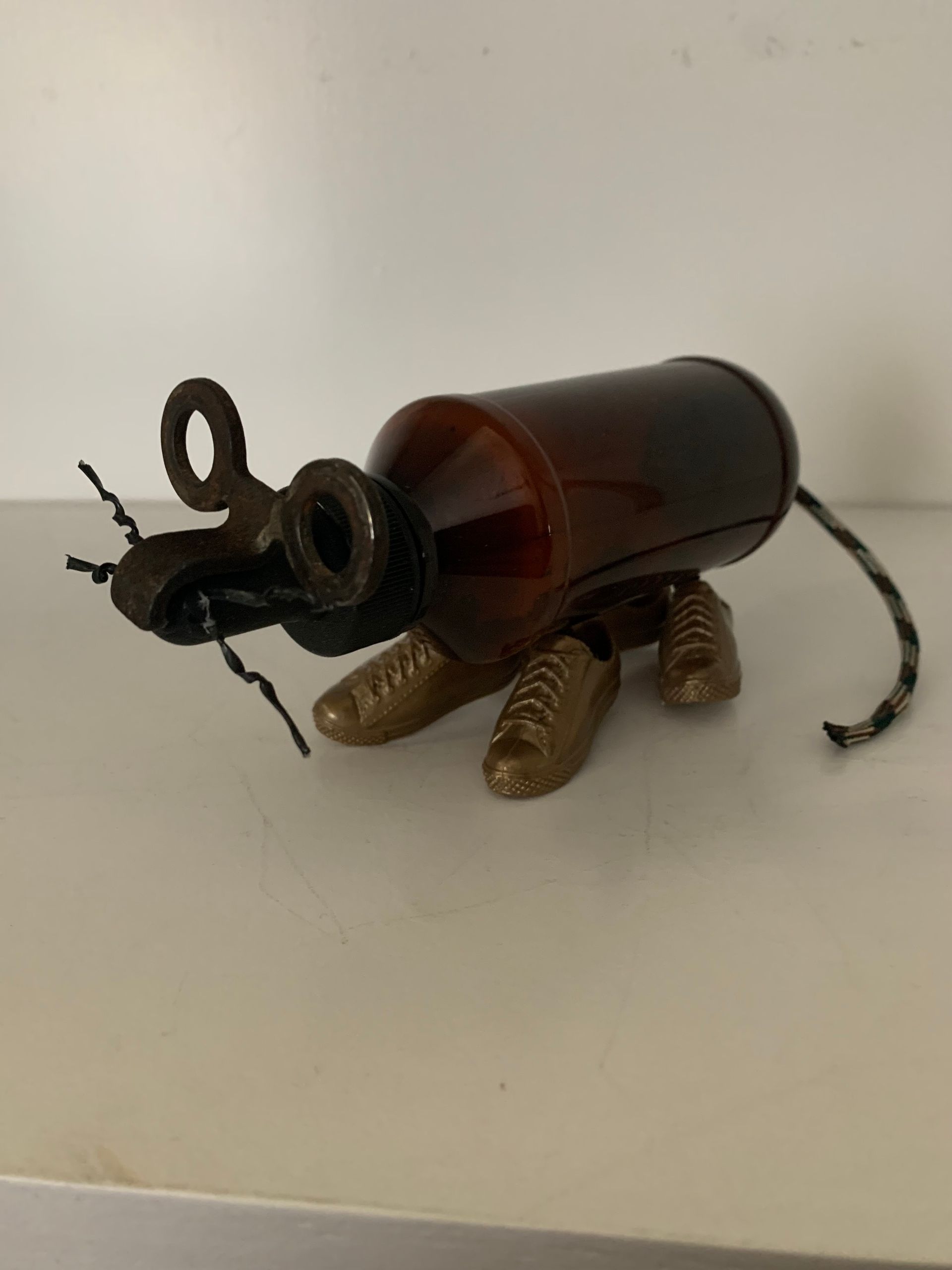 Sculpture of a brown bottle turned into an insect, with metal legs, antennae, and tail on a white surface.  By Mary Berry - Art