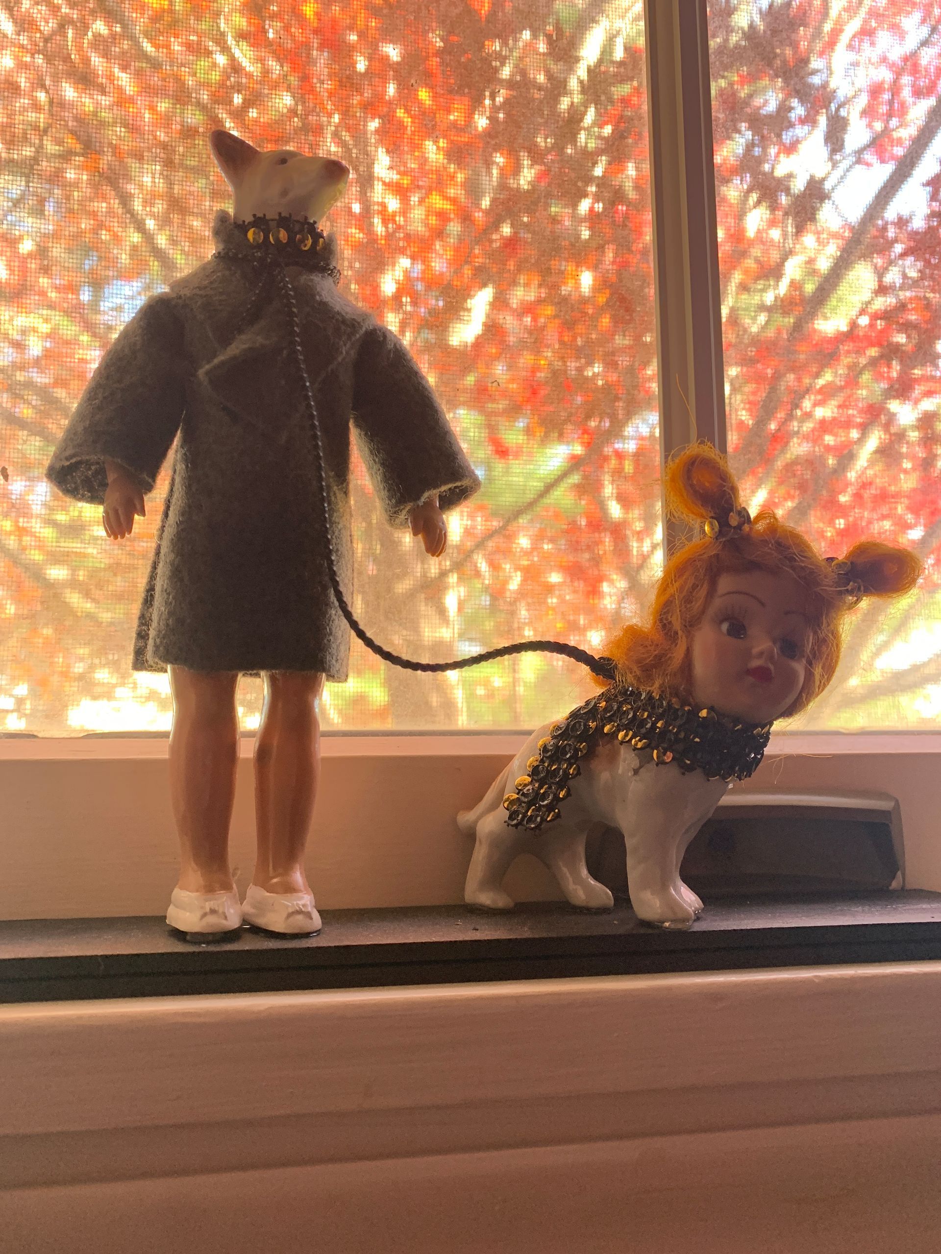 Doll-like figures; one in coat holding leash connected to dog-like figure, on windowsill, fall foliage background.  By Mary Berry - Art