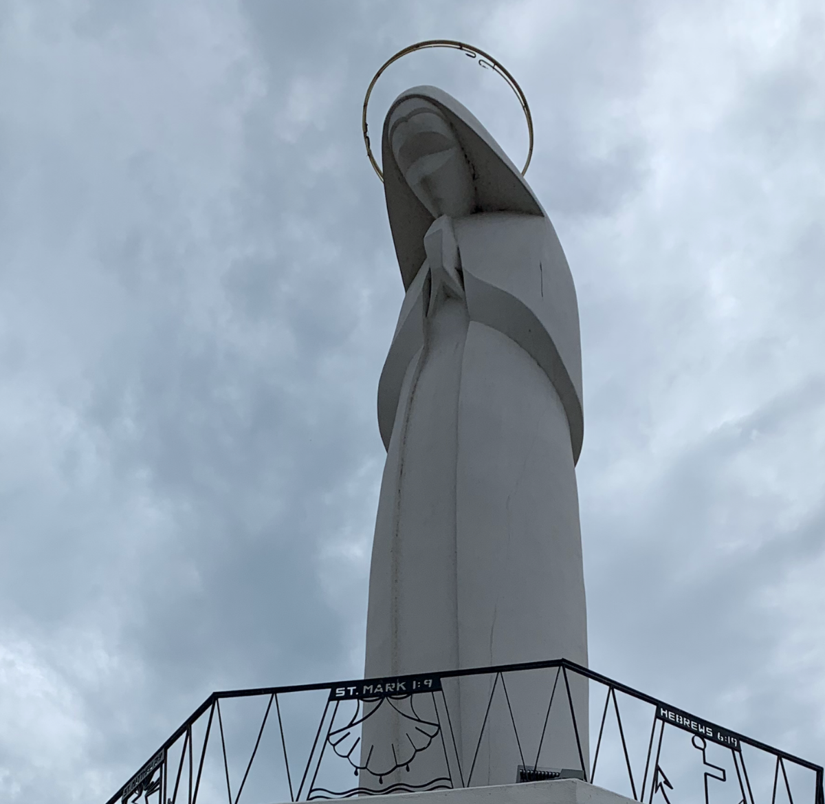 White statue of Our Lady of the River in Grafton, IL  with a halo, arms clasped in front of her, against a cloudy sky.  By Mary Berry - Art