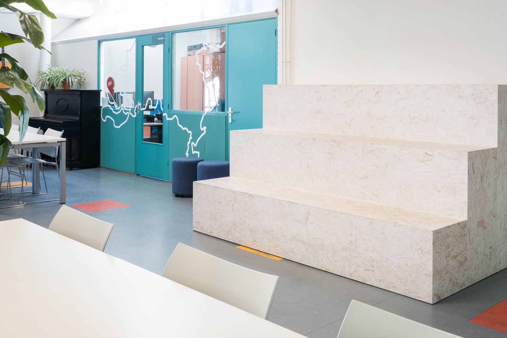 Modern kantoorinterieur met witte, trapsgewijs oplopende zitbanken. Turquoise deuren.