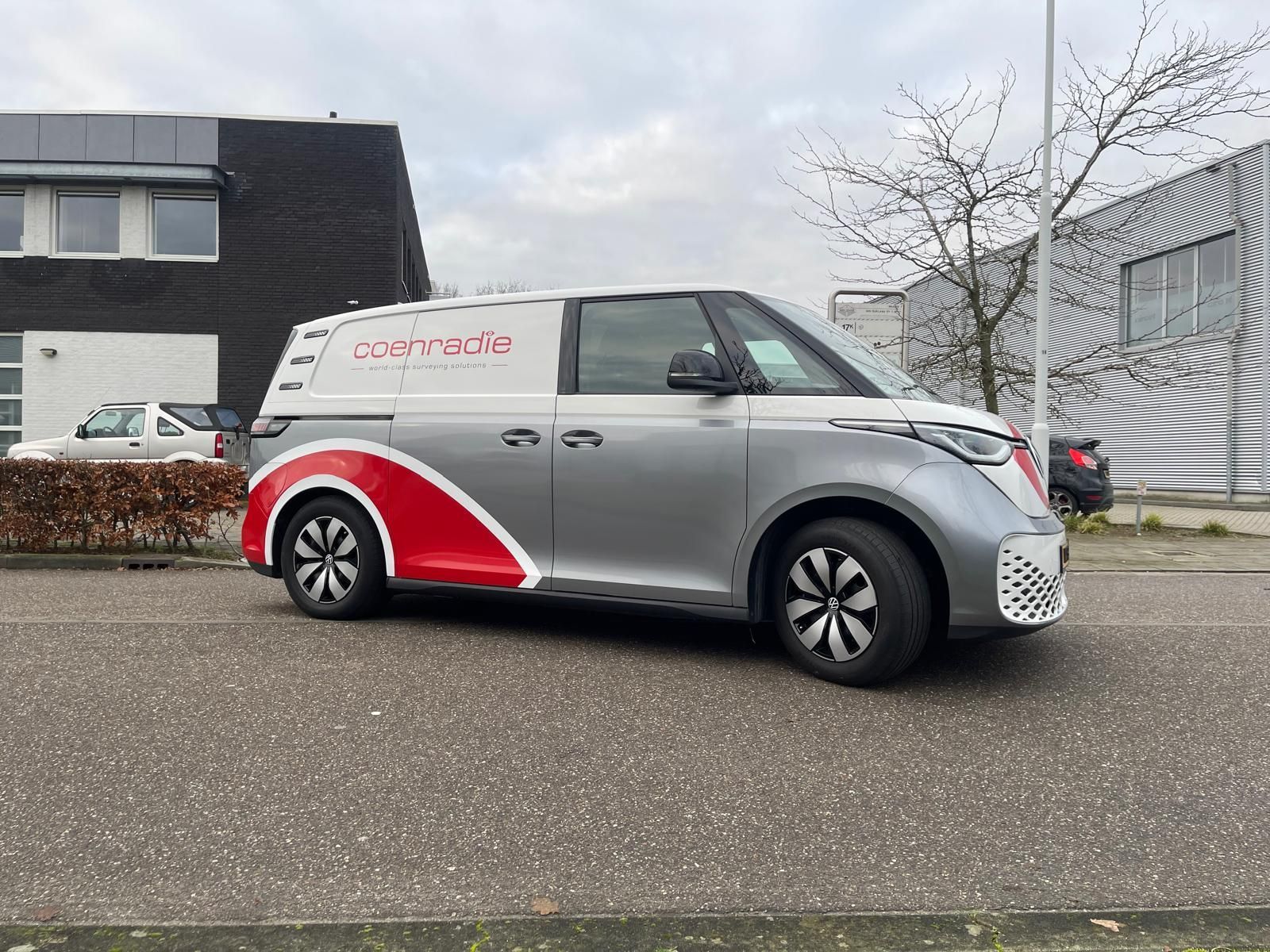 Een zilverkleurige Volkswagen ID. Buzz Cargo bestelbus staat geparkeerd op straat, met rode logo's op de achterwielkasten.