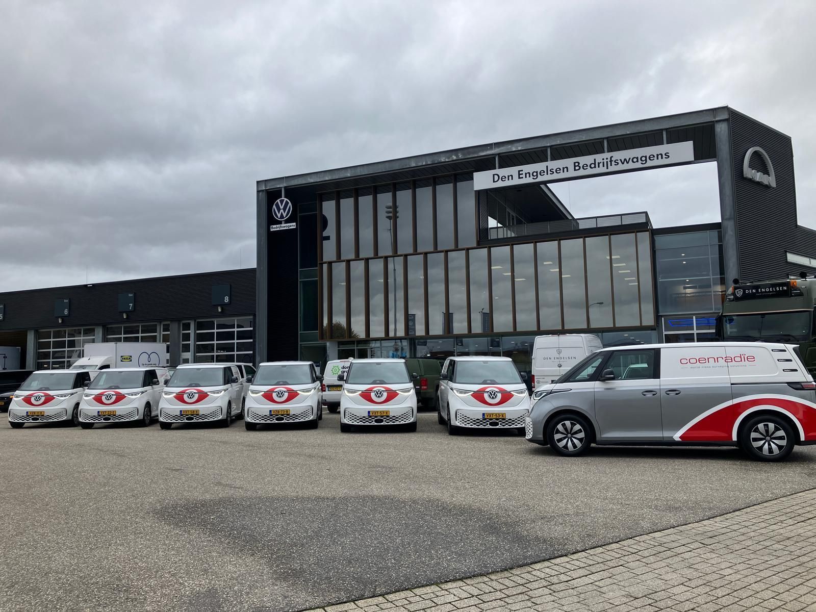 Een rij kleine, witte bestelbusjes met logo staat geparkeerd voor een modern autodealerpand onder een bewolkte hemel.