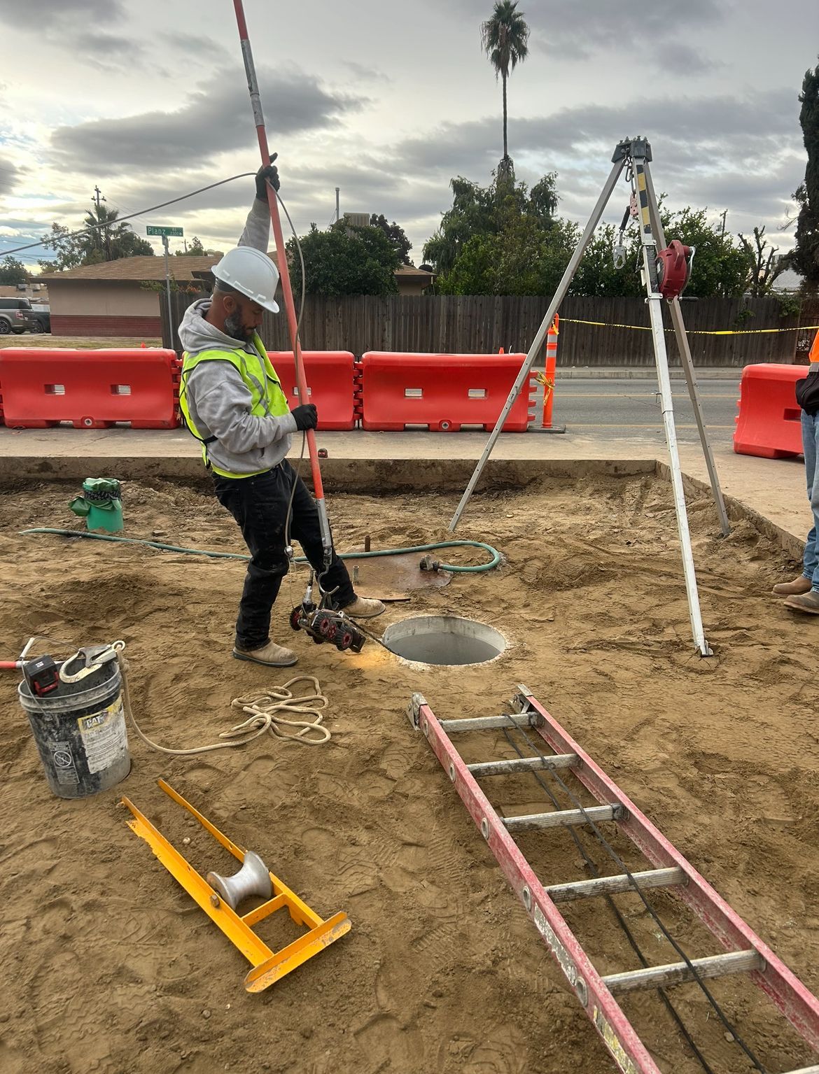 A man in a hard hat is standing in the dirt next to a ladder and an open pipeline doing a video inspection of the pipline.