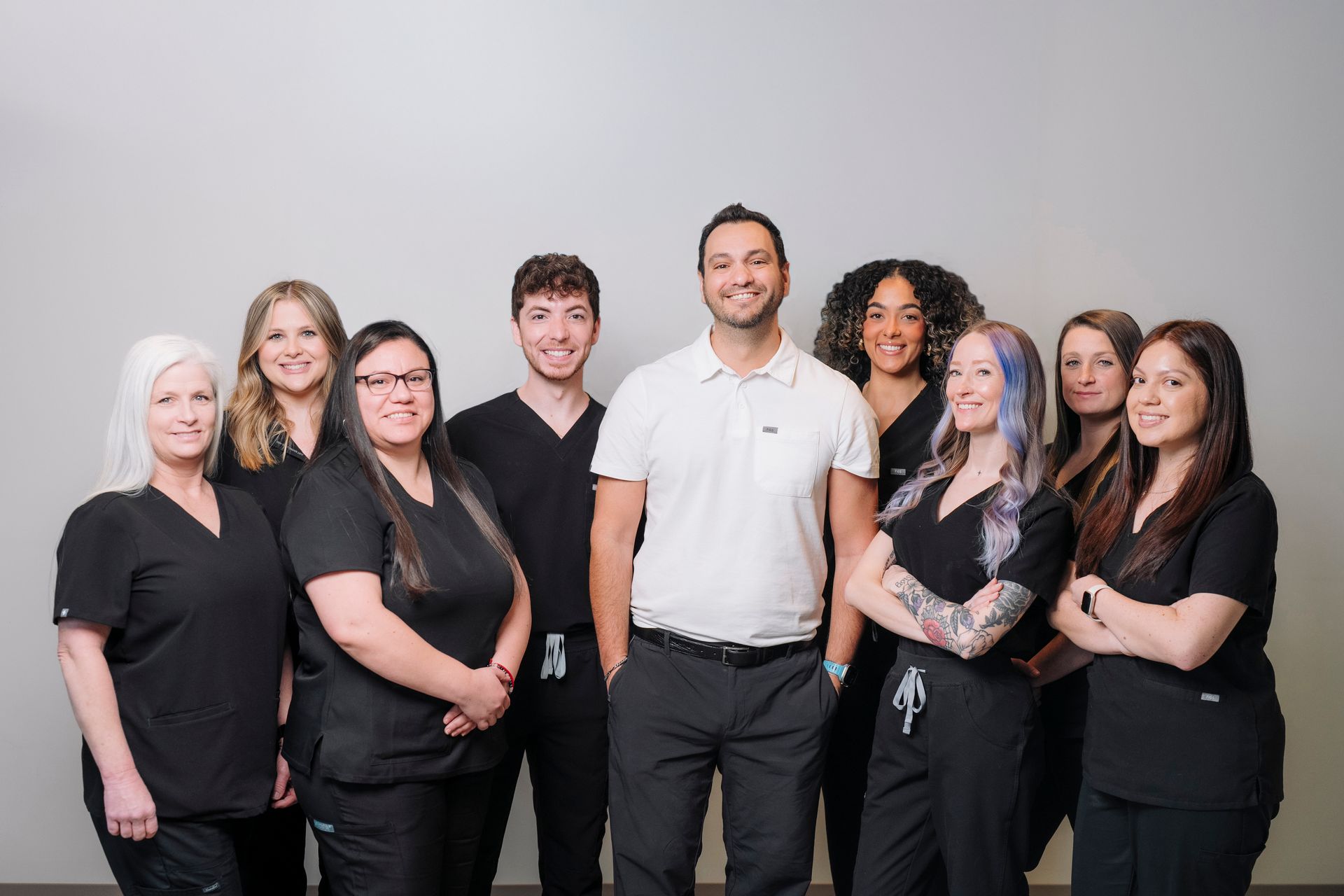 A professional group portrait of nine staff members in black scrubs and one man in a white polo shirt against a gray wall.