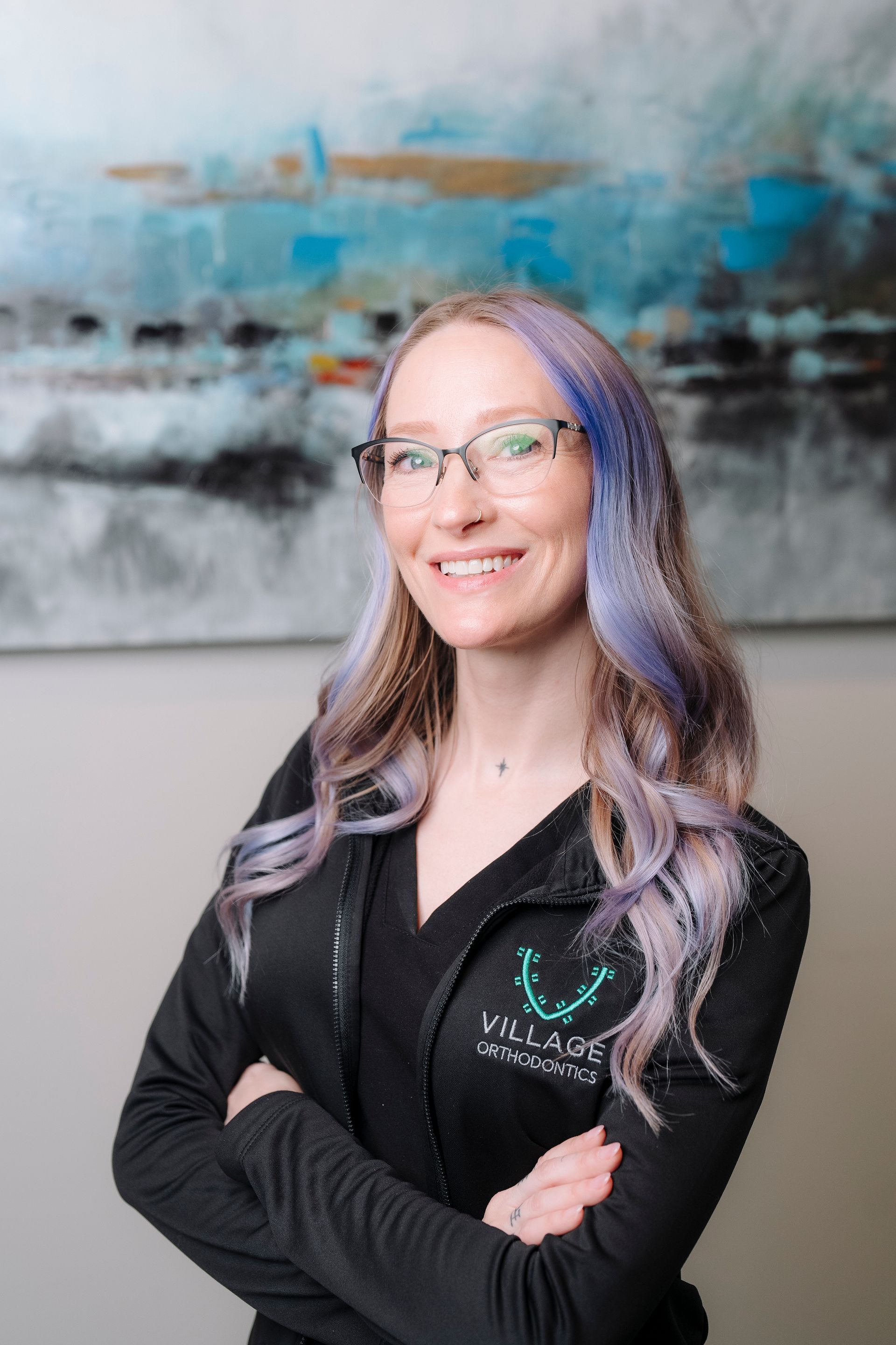 A smiling person with blue-tinted hair and glasses wearing a black jacket with a Village Orthodontics logo, arms crossed.