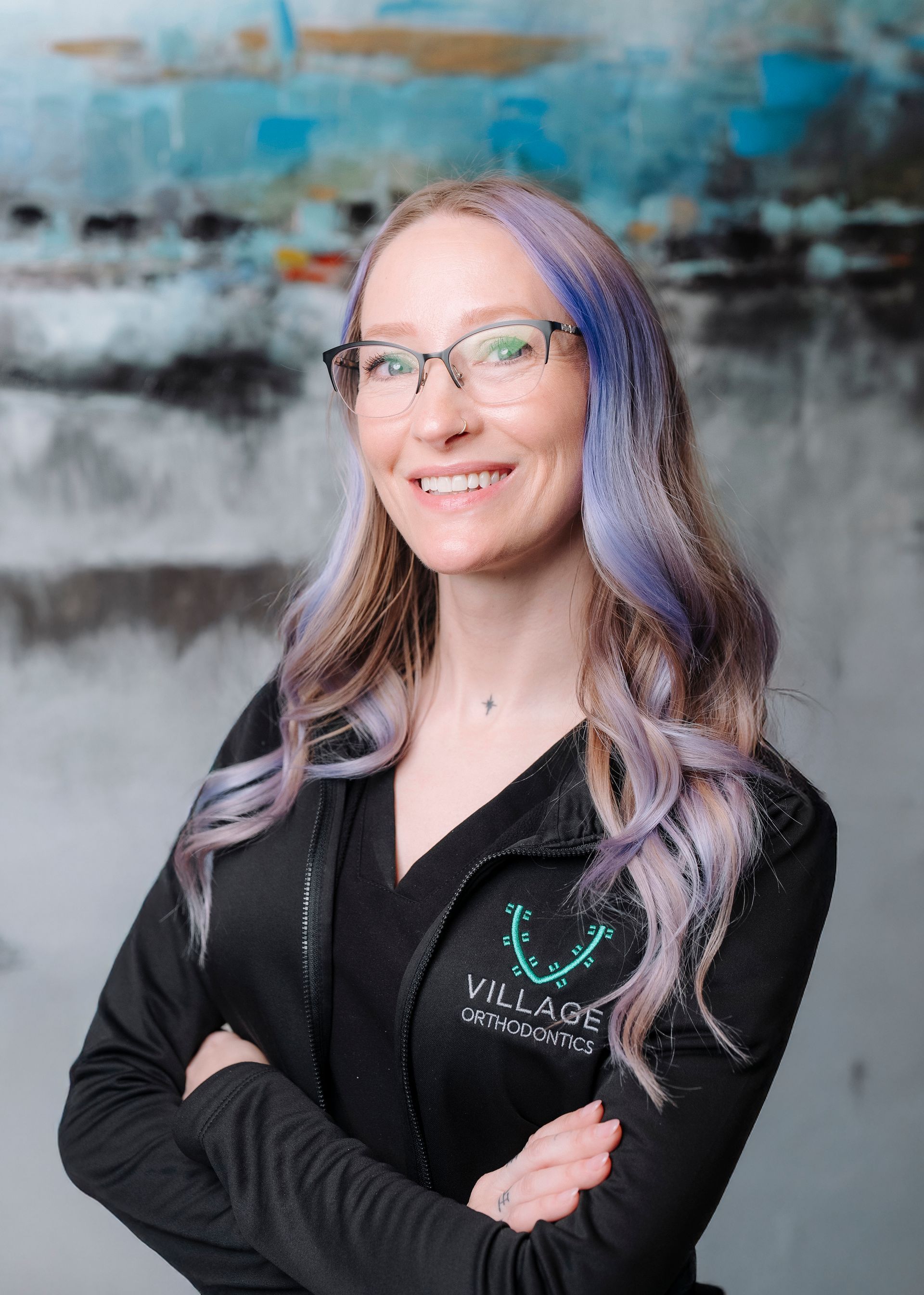 A smiling person with blue-tinted hair and glasses wearing a black jacket with a Village Orthodontics logo, arms crossed.