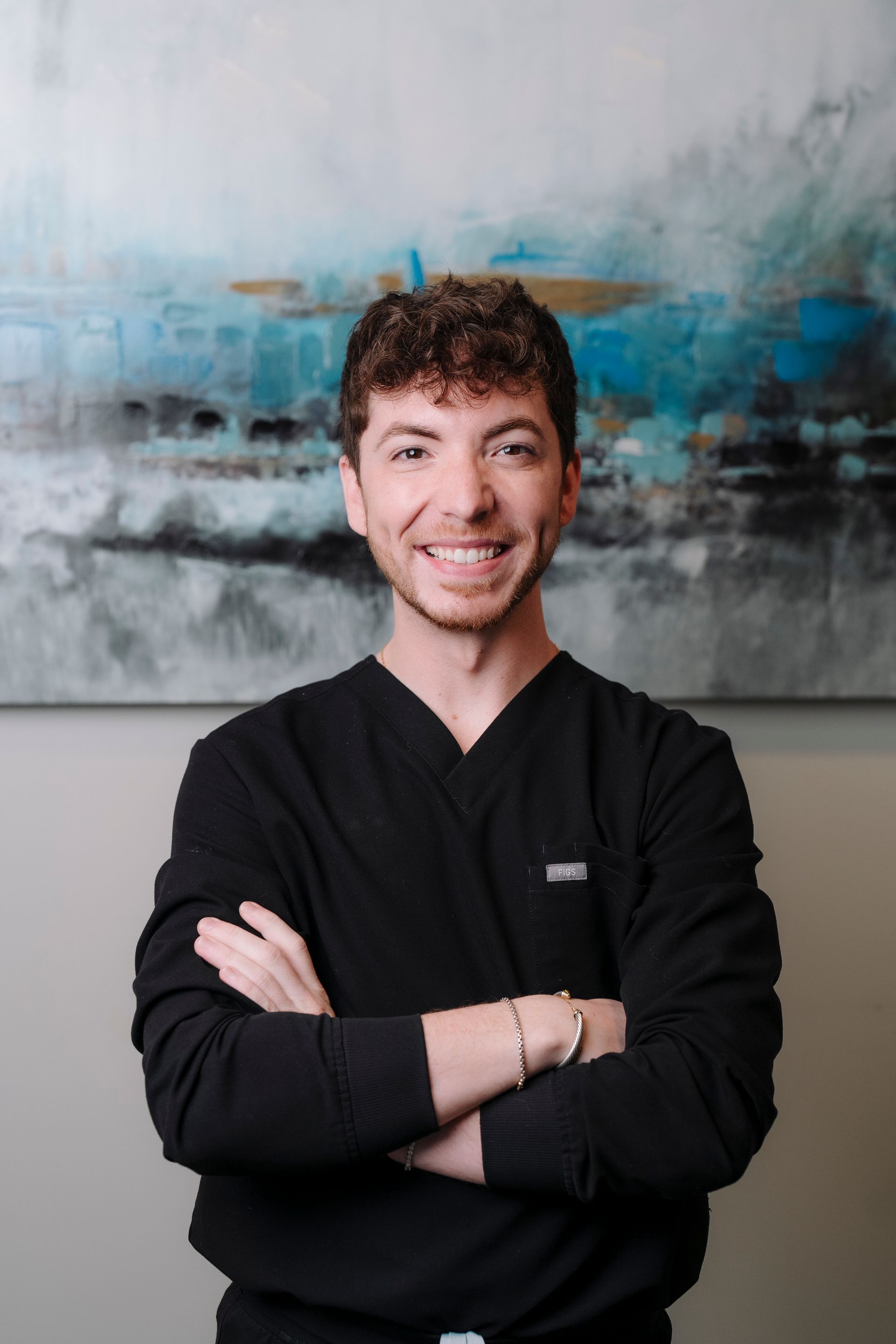A person wearing a black v-neck top with arms crossed, smiling in front of a blue and gray abstract painting.