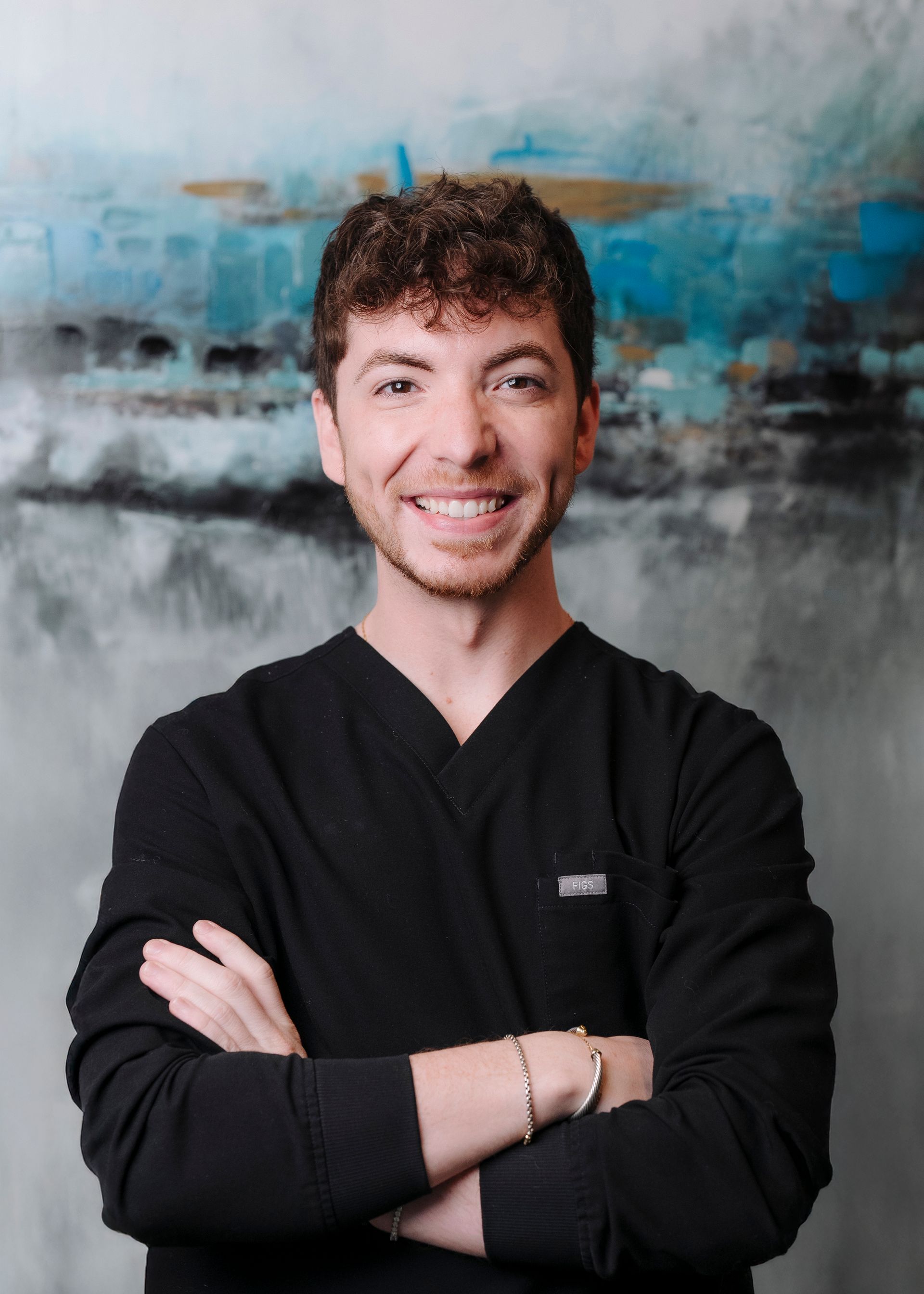 A person wearing a black v-neck top with arms crossed, smiling in front of a blue and gray abstract painting.
