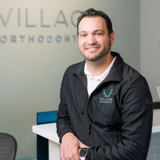 A man in a black quarter-zip sweater with a logo smiles while leaning against a desk in a Village Orthodontics office.