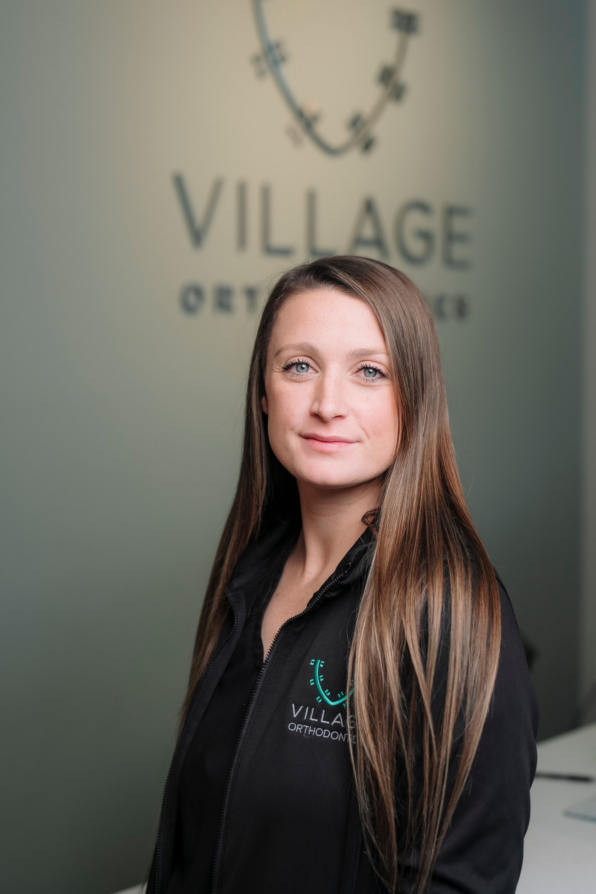 A person with long brown hair, wearing a black top, poses in front of a Village Orthodontics sign on a green wall.