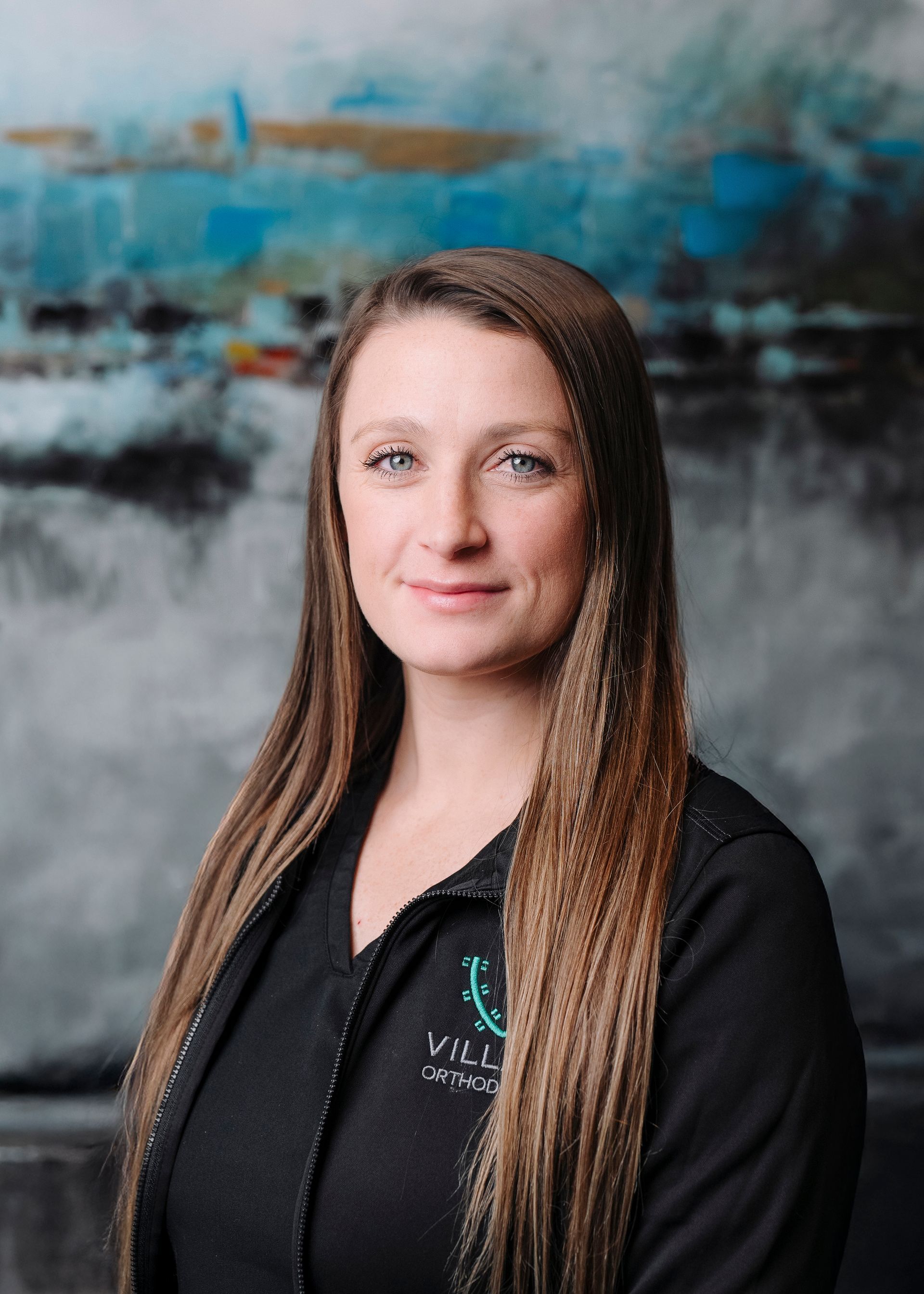 A person with long brown hair, wearing a black top, poses in front of a Village Orthodontics sign on a green wall.