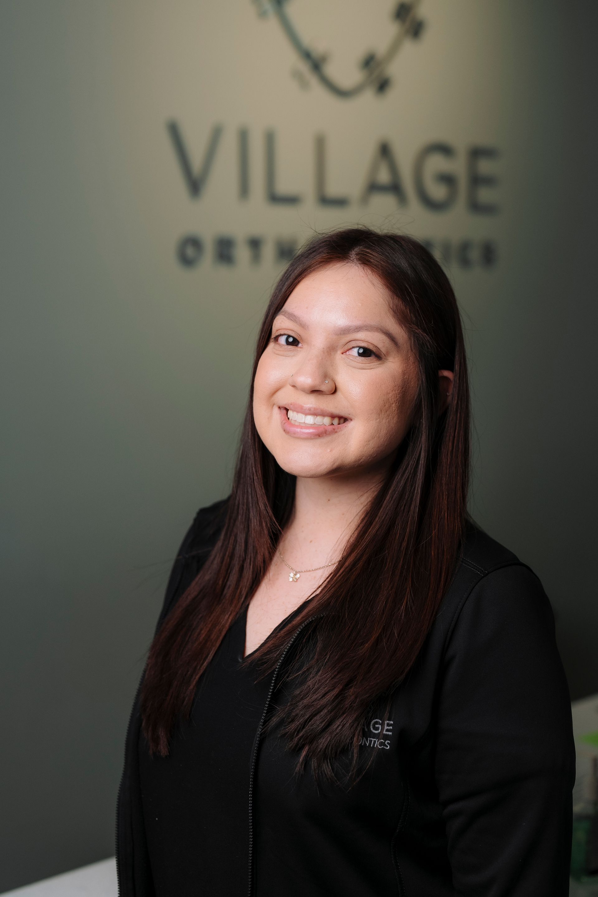 Smiling person in a black shirt standing in front of a Village Orthodontics logo on a dark green wall.