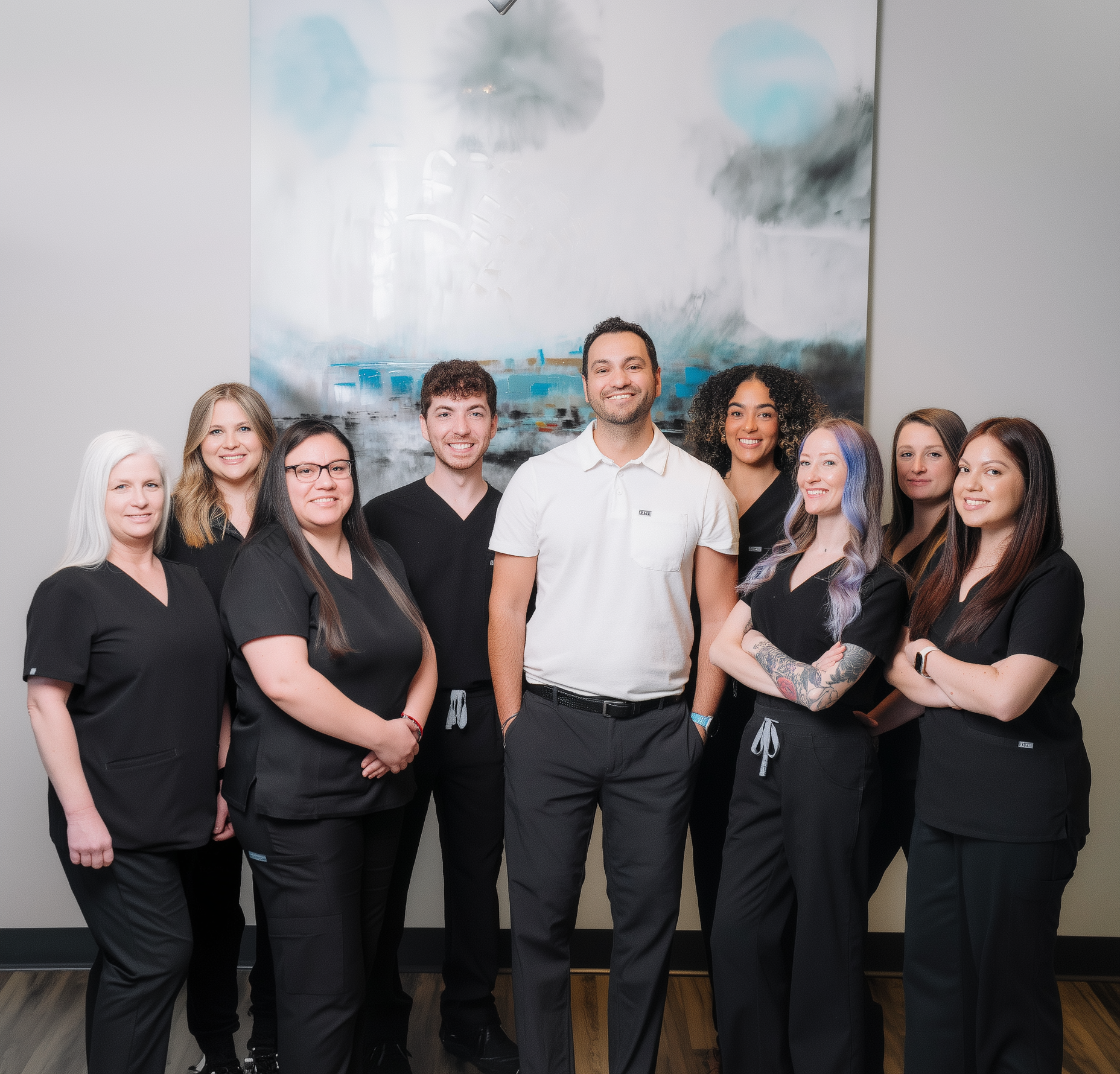 A group of nine professionals in black scrubs and a man in a white polo shirt pose together in a bright office.