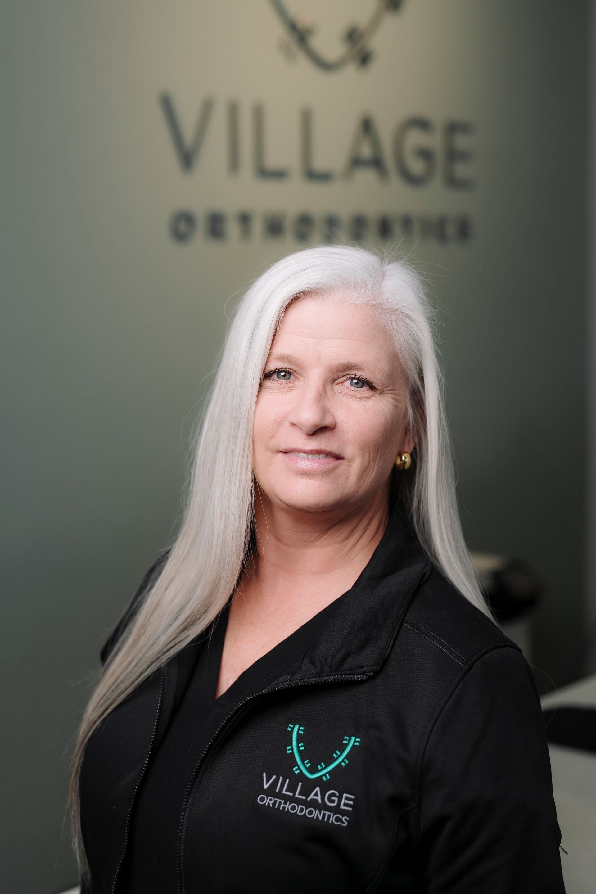 A person with long gray hair wearing a black uniform with a Village Orthodontics logo, standing in front of office signage.