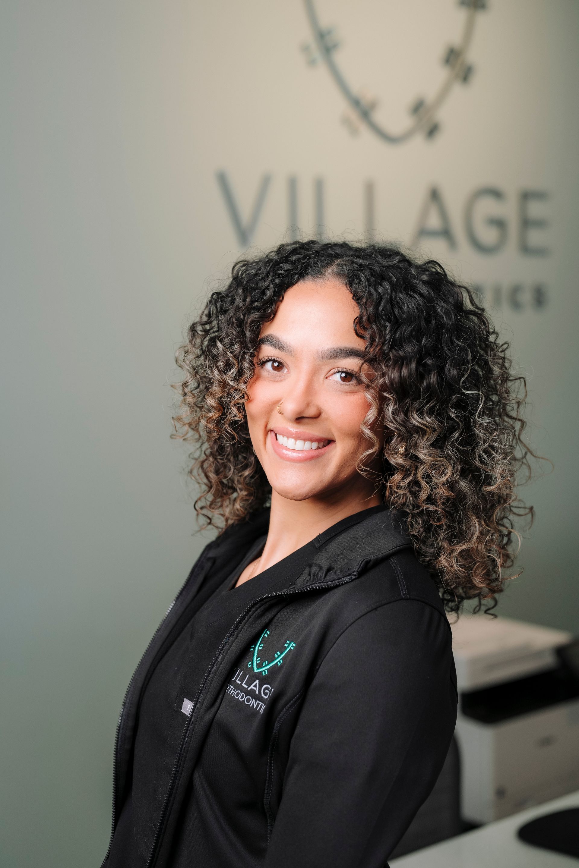 Smiling staff member in black professional attire standing in front of a Village Orthodontics branded wall.