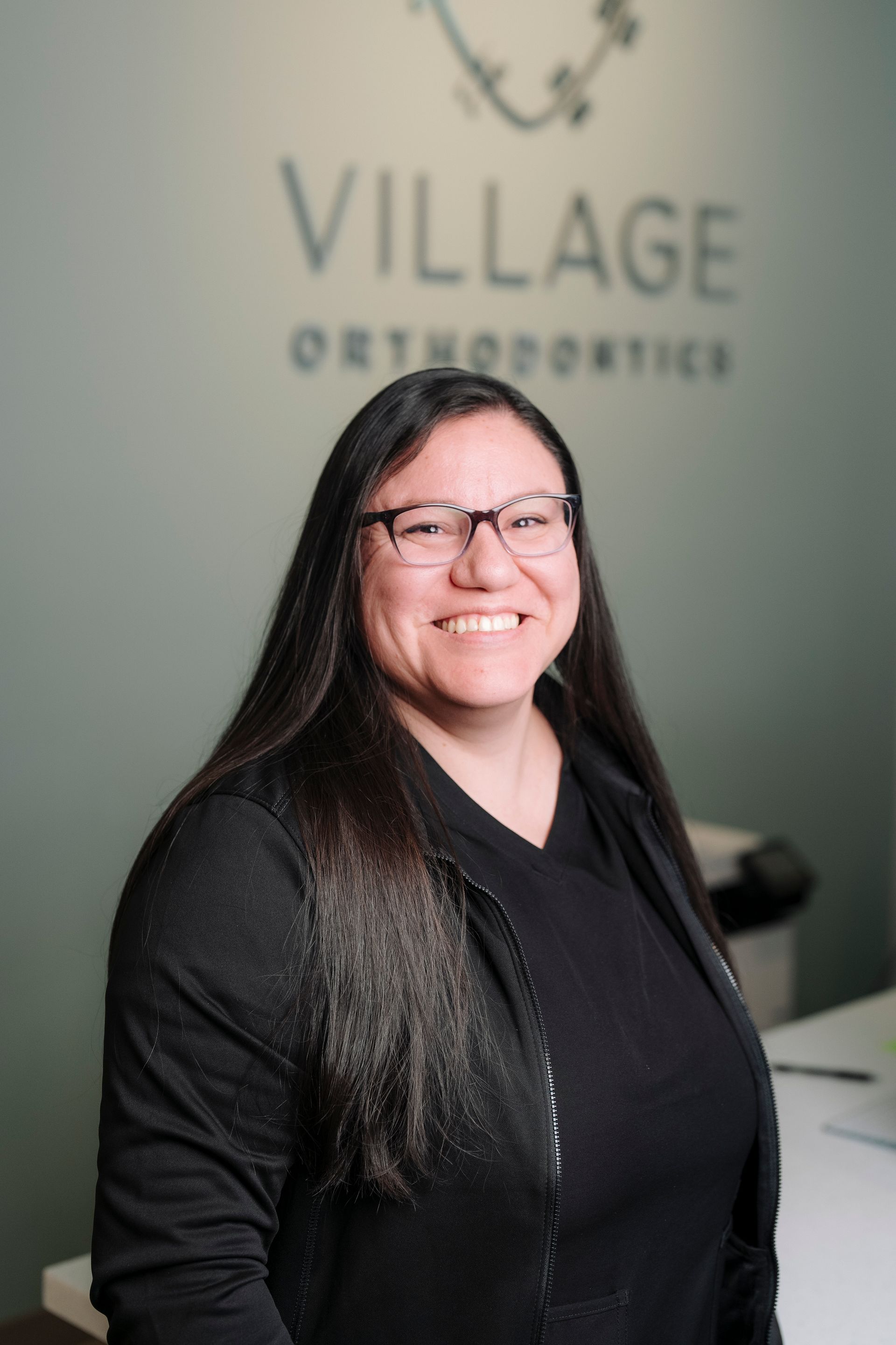 Smiling person with long dark hair and glasses in front of a Village Orthodontics sign.