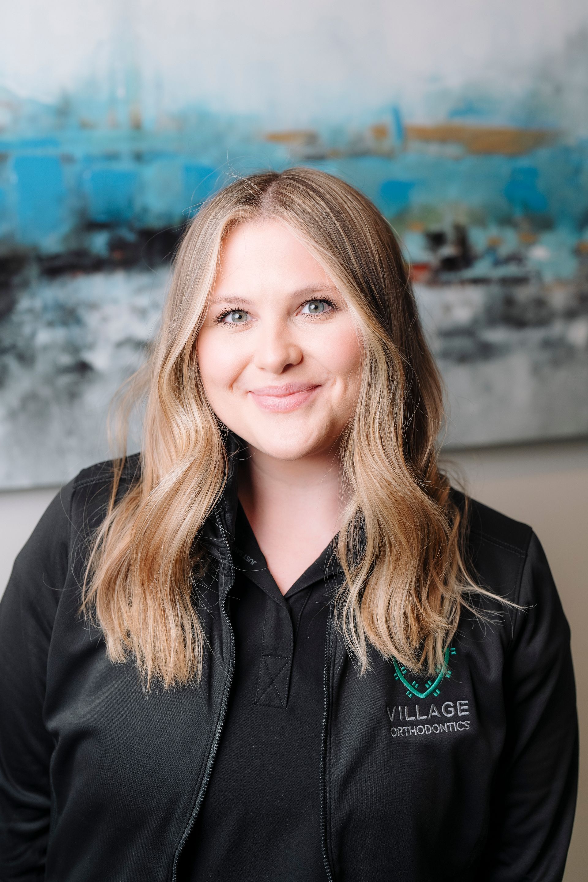 A smiling person wearing a black jacket with a logo, centered in front of an abstract blue and white painting.