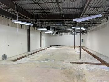 Interior view of a bare commercial space with white walls, exposed ceiling, and floor trenches.