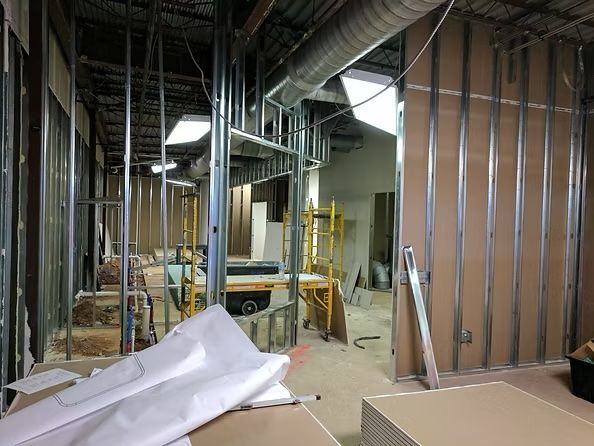 Interior construction site; metal studs and drywall partially installed, scaffolding visible, open doorways, exposed ceiling ductwork.