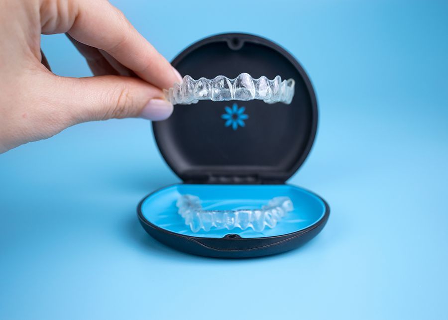 Hand holding clear aligner over a case, against a blue backdrop.