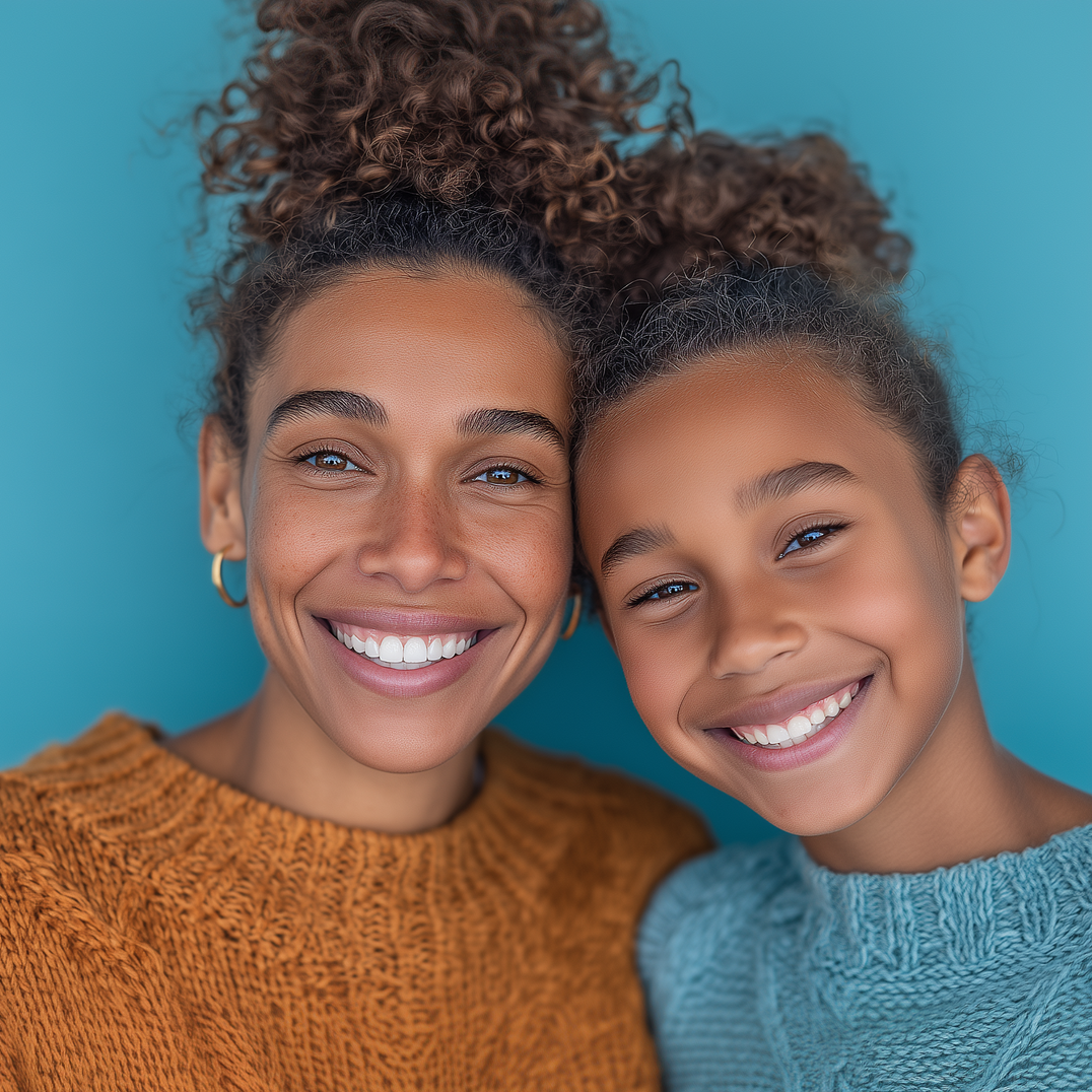 Woman and girl smiling, heads together, against blue background. Woman in orange sweater, girl in blue.