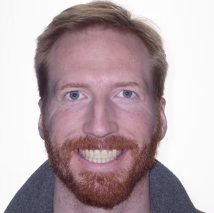 Man with red hair and beard smiling. Wearing a gray jacket.