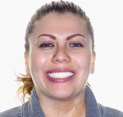 Woman smiling, showing white teeth, wearing grey top. Light skin, brown hair pulled back.