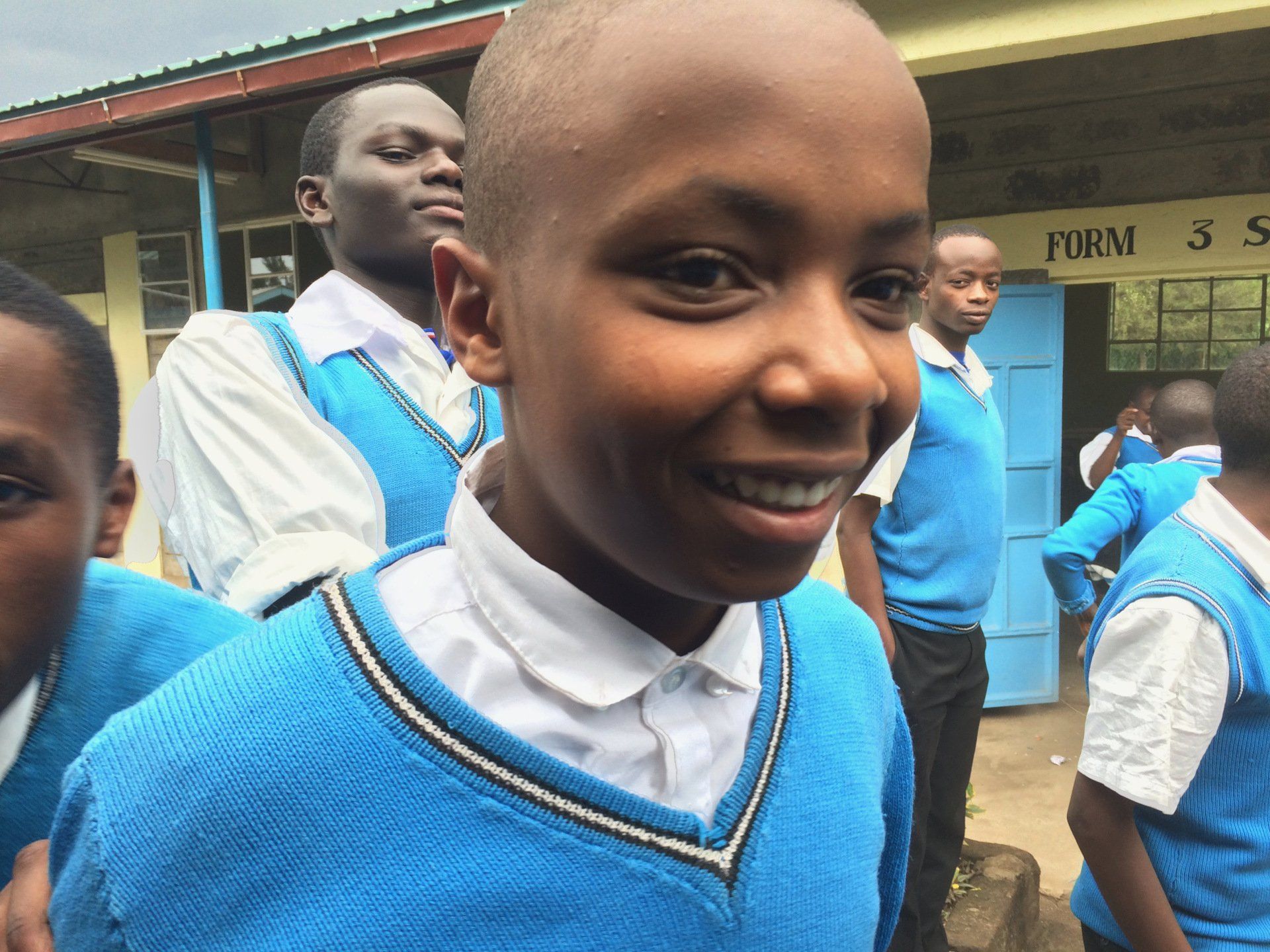 A boy in a blue vest is smiling in front of a sign that says form 3
