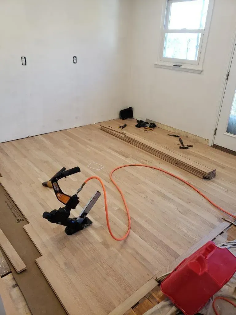 Room with unfinished hardwood floor installation; nail gun, wood planks, tools scattered.