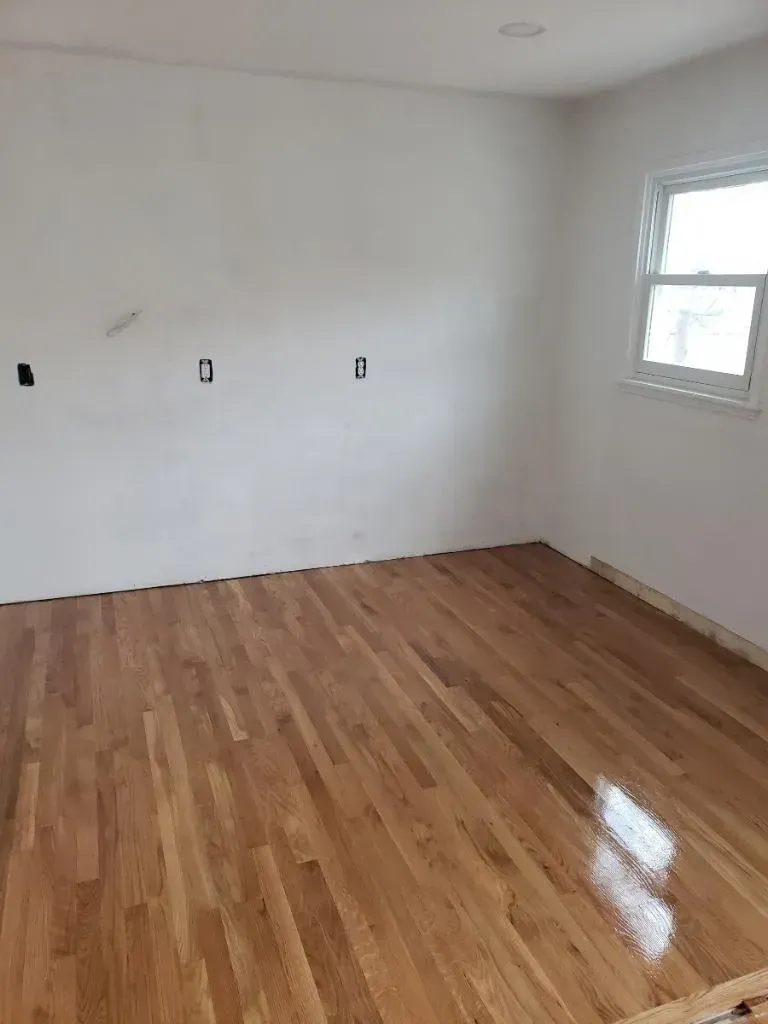 Empty room with hardwood floors, white walls, and a window. Electrical outlets are visible.