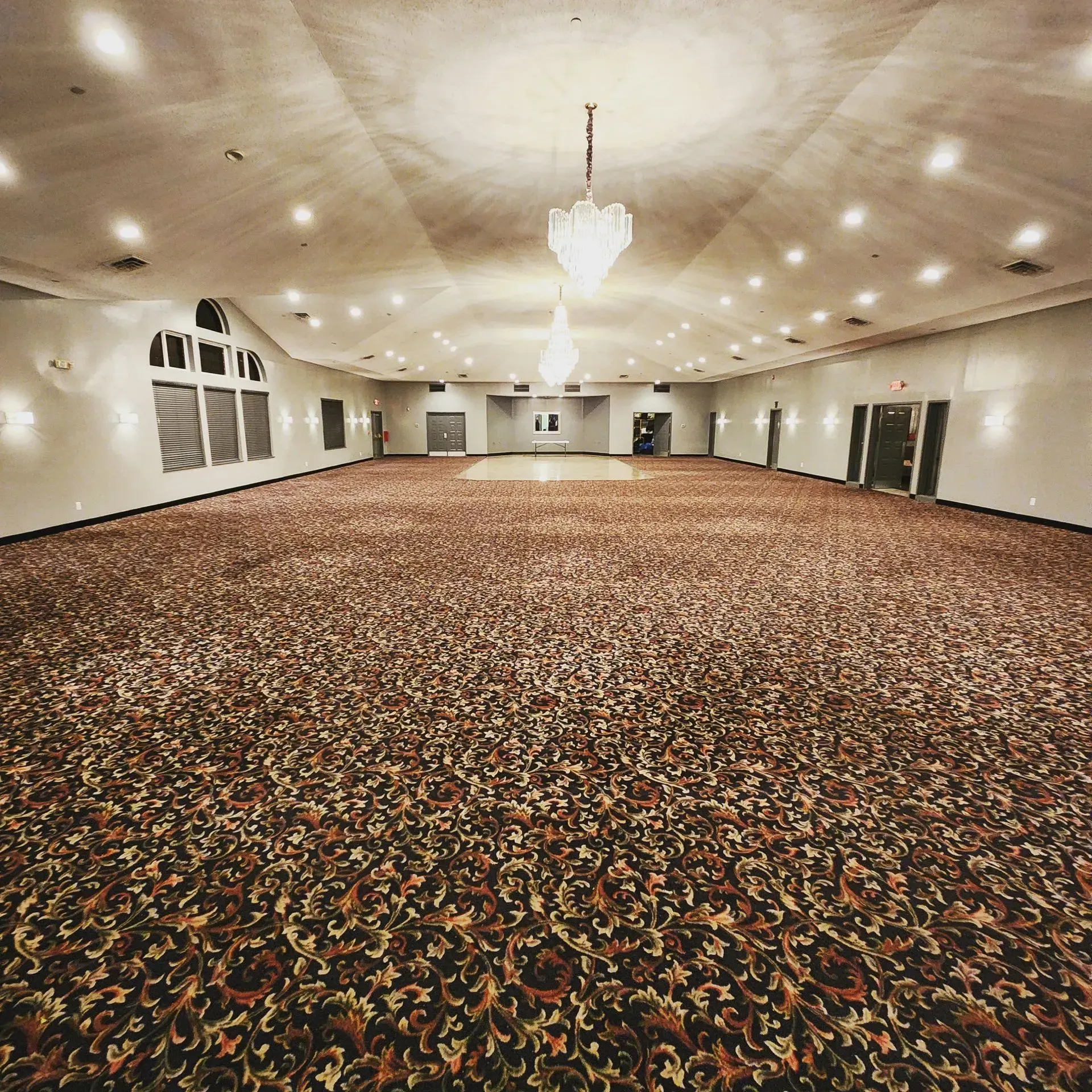 Large, empty ballroom with ornate patterned carpet, chandelier, and wall sconces.