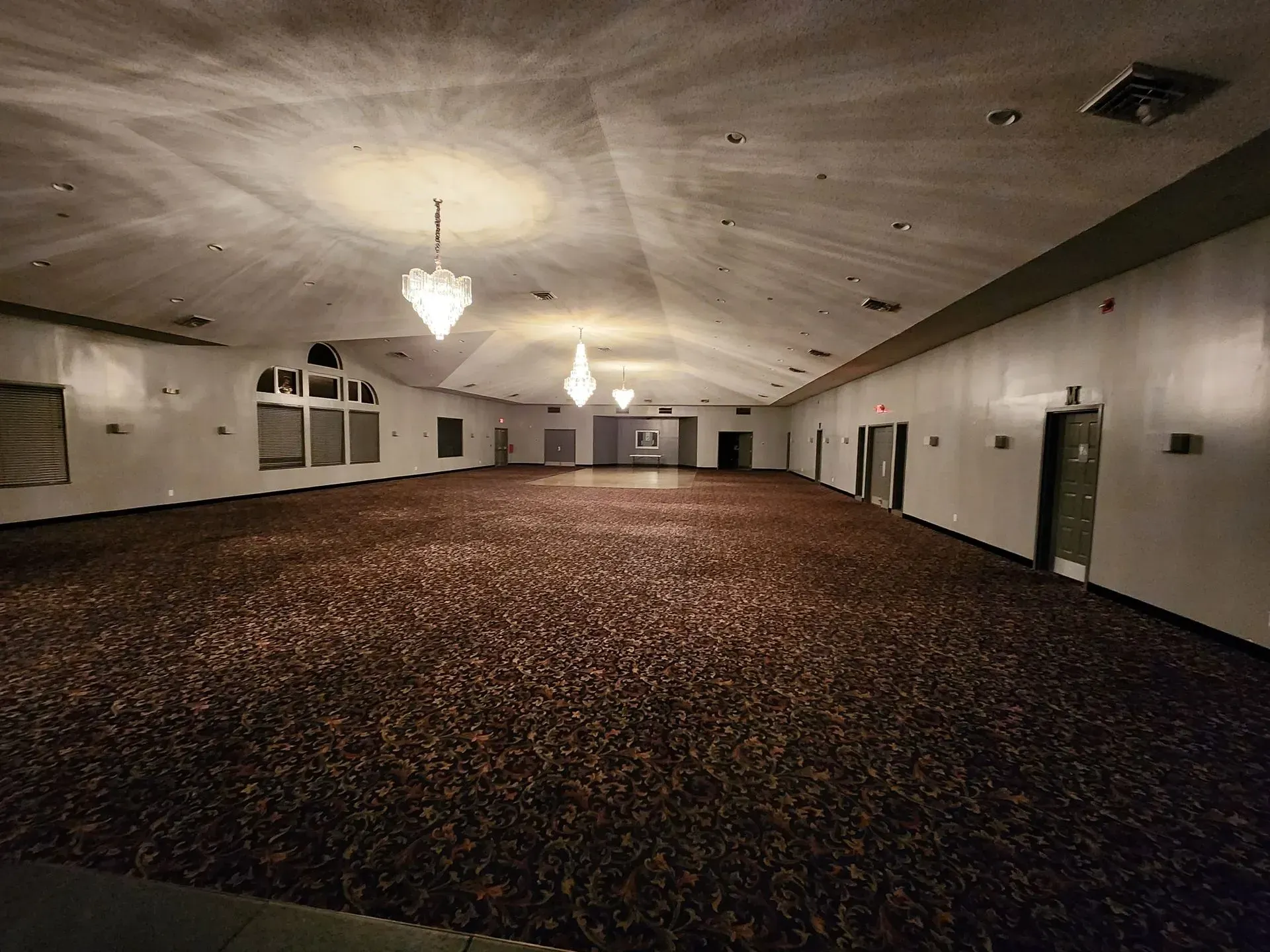 Large, empty ballroom with brown carpet, white walls, and chandeliers.