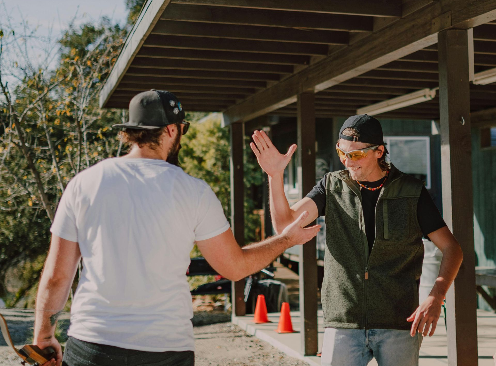 Two men high-fiving outdoors near a wooden structure; one wears a white shirt, the other a vest and sunglasses.